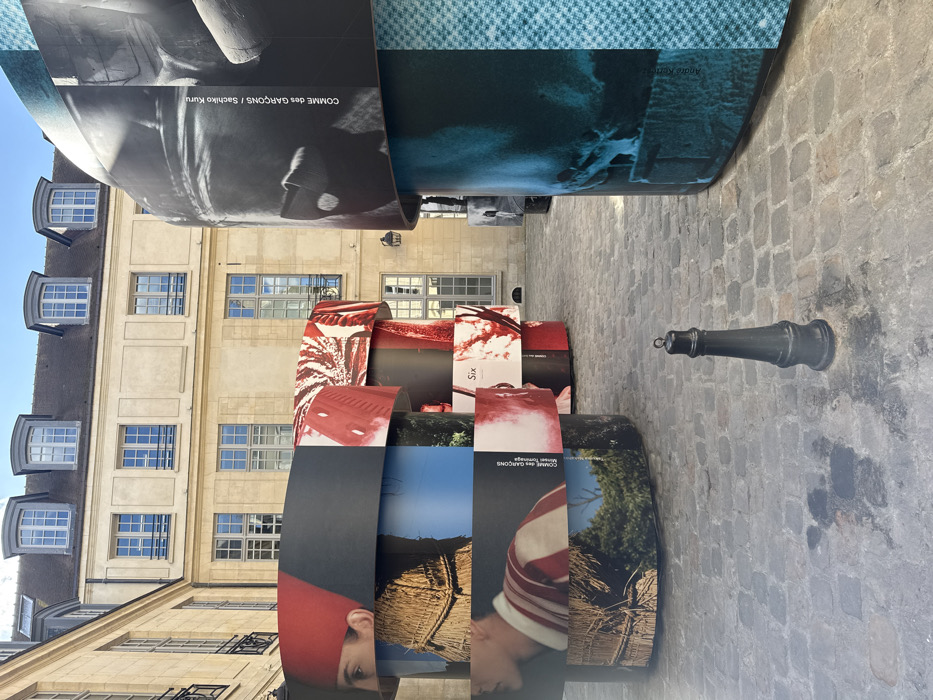 The image depicts an outdoor cobblestone courtyard in Paris, France, bordered by multi-story classical buildings constructed of light-colored stone with numerous rectangular windows and grey mansard roofs featuring dormer windows. The courtyard is primarily occupied by several large, free-standing cylindrical display structures. These cylinders are composed of stacked horizontal segments wrapped with printed photographic and graphic imagery.
Visible imagery on the cylinders includes a person wearing a red head covering, a person in a red and white striped top, sections of straw-like material, green foliage against a blue sky, and abstract black-and-white and teal-toned designs. Text displayed on the structures identifies "COMME des GARÇONS / SIX" and "Sachiko Kuru."
In the foreground, a single black, conical metal bollard with a metal loop at its top stands on the cobblestone ground. The ground exhibits various dark stains and shadows, indicating direct sunlight from above and to the left. No individuals are present within the frame.