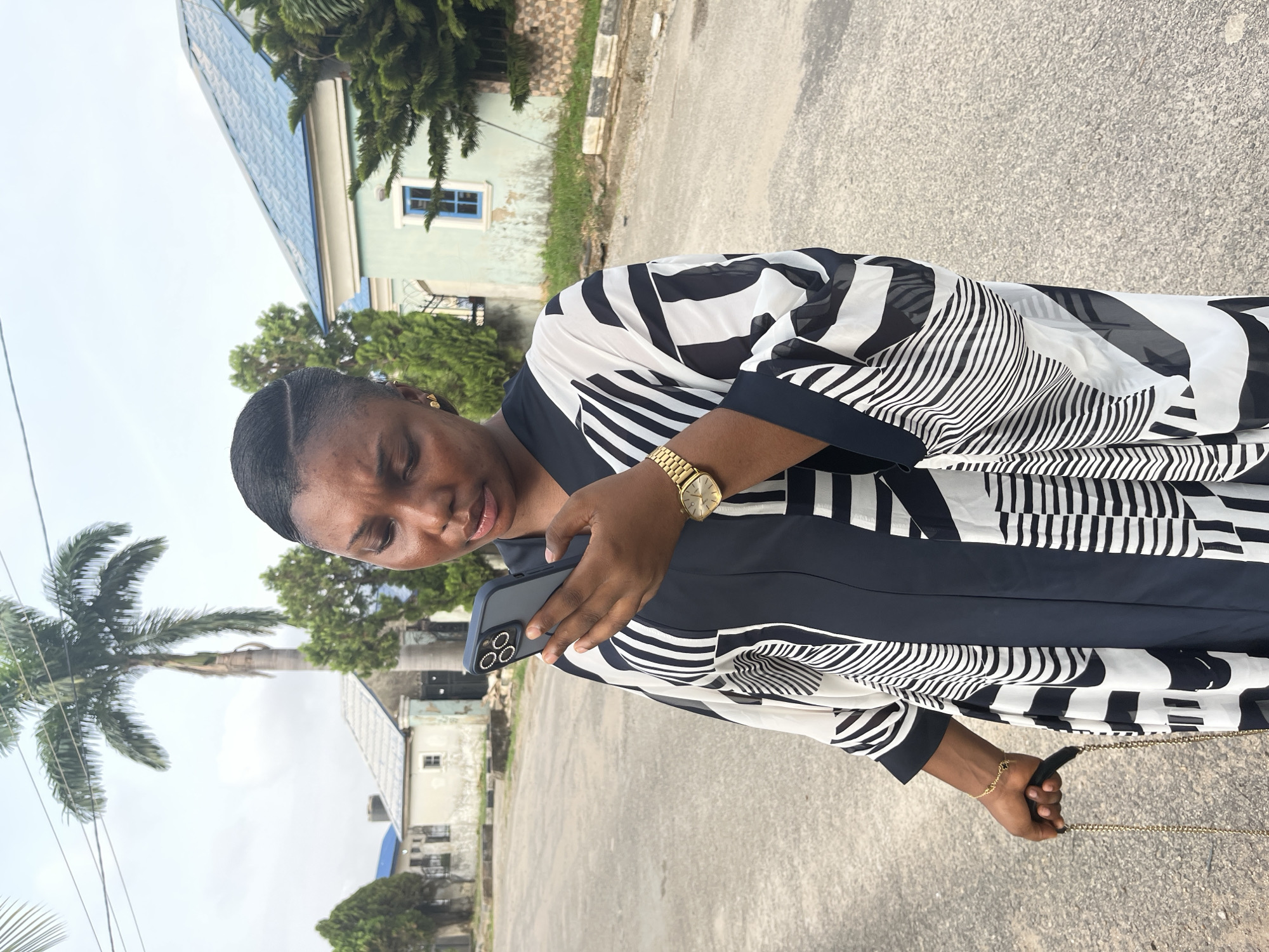 A woman is standing on a paved road, looking at a smartphone she holds in her hands. She is wearing a black and white patterned dress and a gold watch. A palm tree is to her left. Behind her are buildings, including one with a blue roof. The location may be in Abuja, Nigeria.