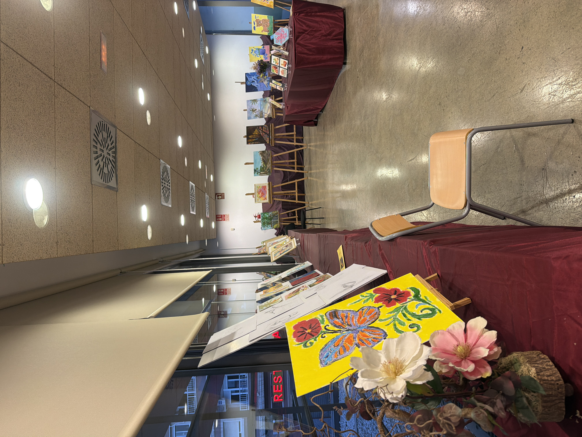 This image depicts an art exhibition, likely in a public space or community center, in Elche, Spain. The scene is set indoors with a polished concrete floor reflecting the overhead lighting. Large windows line the left side of the room, revealing glimpses of an urban environment with buildings and signage, including the word "REST" in red letters on a facade.
The exhibition features numerous paintings displayed on easels and tables. In the foreground, a table covered with a dark red tablecloth displays several artworks. A bright yellow canvas with a painting of a butterfly and red flowers is prominently positioned on a small easel. To its left, a floral arrangement with white and pink flowers is placed in a decorative base.
Further into the room, more paintings are arranged on easels. Some are landscapes, while others appear to be portraits or still lifes. A large table also covered in a dark red tablecloth is set up in the mid-ground, showcasing smaller framed artworks and possibly other art supplies.
A simple, light-colored chair with a metal frame is placed in the foreground, facing the artworks, suggesting a viewing area for visitors. The ceiling features acoustic tiles and multiple circular light fixtures, along with ventilation grates. The overall impression is of an organized and well-lit display of local art. No people are visible in the image.