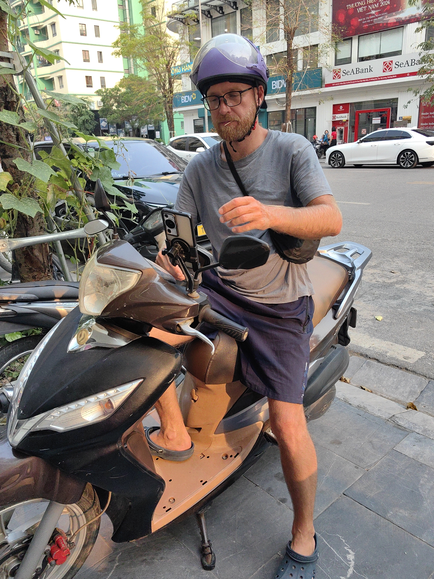 An eye-level, full shot features a Caucasian male seated on a brown scooter parked on a street in Hong Gai Ward, Vietnam. The man is wearing a grey t-shirt, navy shorts, dark blue Crocs, glasses, and a purple helmet. He holds a phone attached to the scooter's handlebar.
The background includes a sidewalk, street, buildings, parked cars, and street trees. A bank with red signage is visible. The scooter is parked on a grey tile sidewalk. The street is asphalt, and a white car is parked further down the road. The buildings are light-colored with green trim. Vietnamese bank signs are legible.
The lighting is bright and natural, suggesting a sunny day.