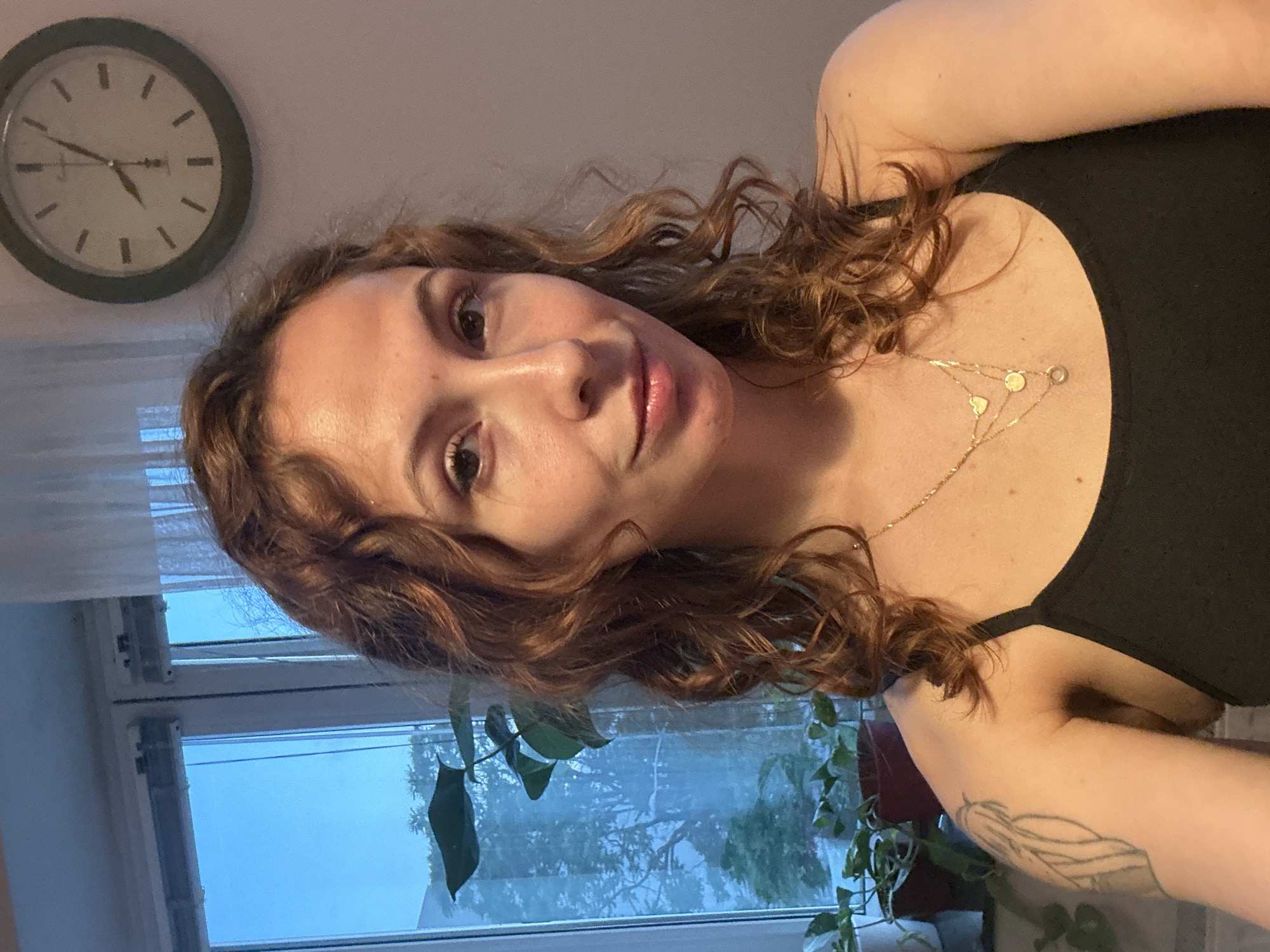A woman with curly brown hair and fair skin is posing for a selfie. She is wearing a black top and a gold necklace with layered pendants. A tattoo is visible on her left arm. Behind her is a window with sheer curtains, a view of trees, and daylight indicating an outdoor scene. A green, round clock hangs on the wall to the left of the woman, displaying a time of approximately 4:50. Indoor plants are visible near the window. There is no explicit information to determine if the location is Novi Sad, Serbia.