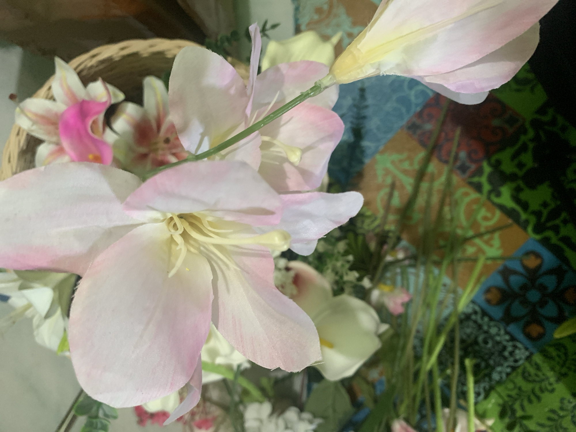 The image shows a close-up of artificial flowers in a basket, with a colorful patterned fabric underneath. The flowers are predominantly light pink and white with some yellow accents. The basket appears to be made of woven material. The fabric has a patchwork design with geometric patterns in shades of blue, green, and brown. The overall scene suggests an indoor setting, possibly a home or a decorative display. There are no people or animals visible in the image. The lighting is soft, and the focus is on the floral arrangement. The image does not provide any information about the city of Kuala Pembuang, Indonesia.