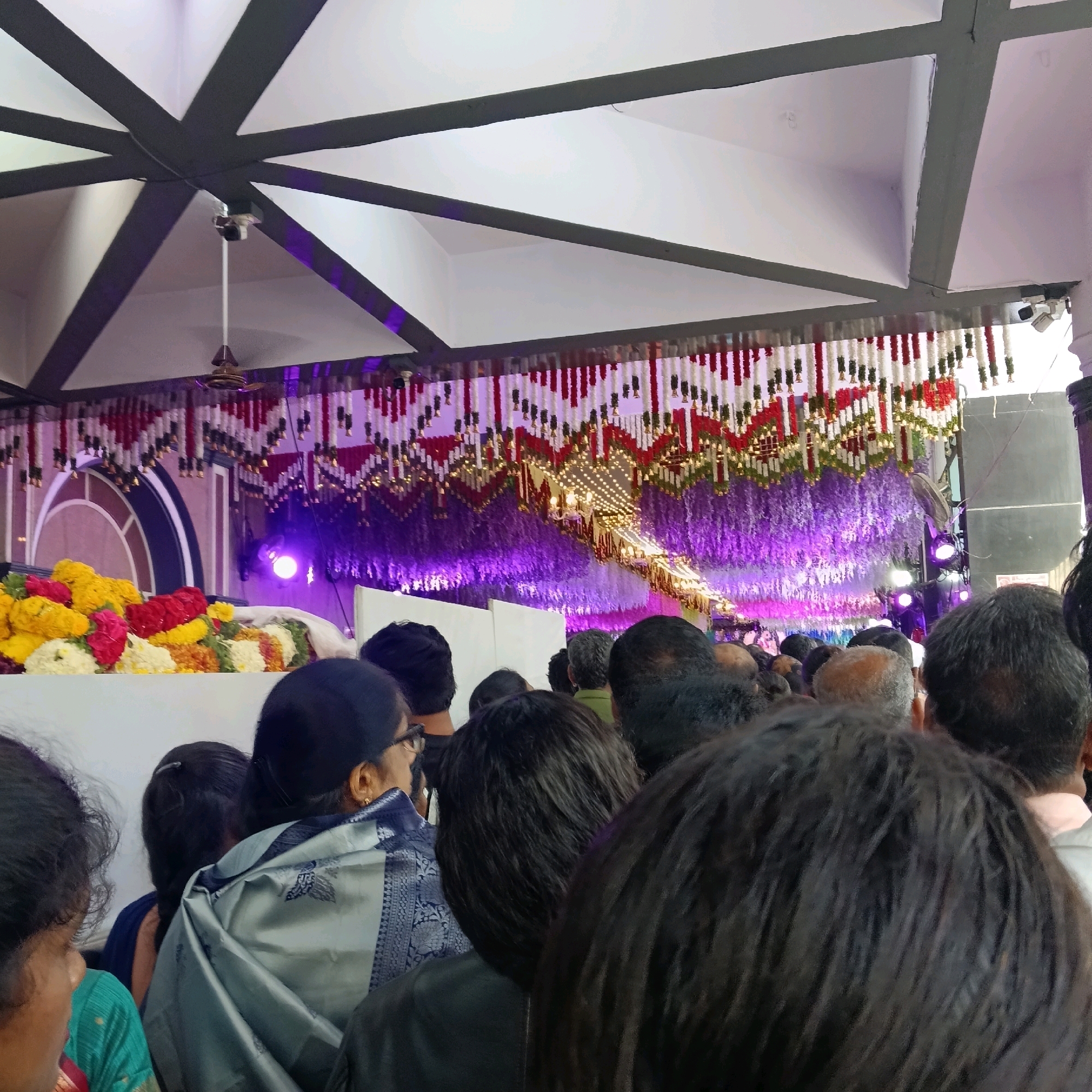 The image shows a gathering of people indoors, likely at an event or ceremony. The scene is decorated with floral arrangements, specifically a large display of yellow, red, and orange flowers on a white surface in the foreground. Above the flowers, there is a ceiling adorned with red and white garlands and a cascade of purple decorations, illuminated by purple lighting. Several people are visible, mostly their backs and heads, indicating they are facing a central point or stage not fully in view. The ceiling has a geometric design with dark beams against a white background. A ceiling fan is visible in the upper left portion of the image. The setting appears to be a formal or celebratory indoor venue in Bengaluru, India.
