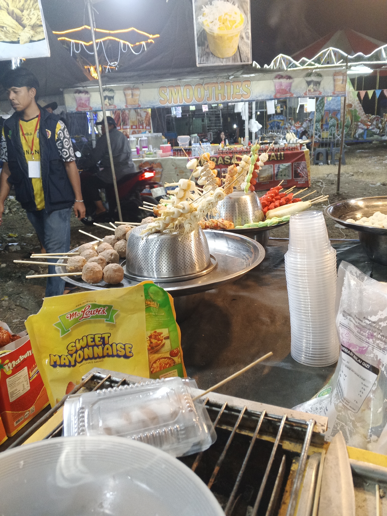 This image depicts a night-time scene at an outdoor food stall, likely a street market or fair, in Sukabumi, Indonesia. In the foreground, a selection of food on skewers is displayed, including meatballs, seafood, and what appear to be sausages. A yellow packet of "Sweet Mayonnaise" is prominently placed in front of the skewers. To the right, a stack of clear plastic cups is visible, along with a partially obscured bag containing nutritional information in Indonesian.
In the mid-ground, a man wearing a dark vest over a yellow shirt with black and white patterned sleeves walks away from the camera, appearing to be an employee or vendor. Behind him, a motorcycle is parked. Further in the background, a stall with "SMOOTHIES" signage is visible, along with other food stalls and a colorful carousel or similar amusement ride. The overall environment suggests a lively, open-air gathering. The ground is uneven and appears to be dirt or gravel. The lighting is dim, with string lights and stall illumination creating a festive atmosphere.
