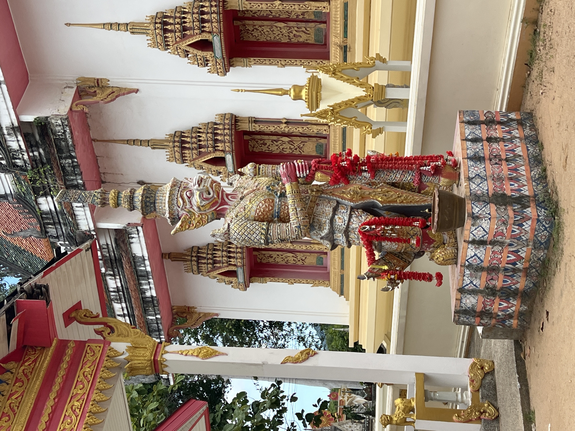 This is a medium shot, eye-level, outdoor photograph of a statue in front of a white building with red and gold trim. The statue is of a large, humanoid figure with a red face, intricate gold armor, and a tall, ornate headdress. Red garlands are draped around the statue's neck and arms. The statue stands on a decorative pedestal with a patterned base. Behind the statue, the building features several windows with gold trim and red interiors. The roof of the building is tiled and appears weathered, with some greenery growing from it. The ground in front of the statue is sandy and there are some fallen leaves. To the left of the statue, a white pillar with gold decorative elements is visible. The overall scene suggests a religious or cultural site. The location is identified as Baan Harn, Thailand.