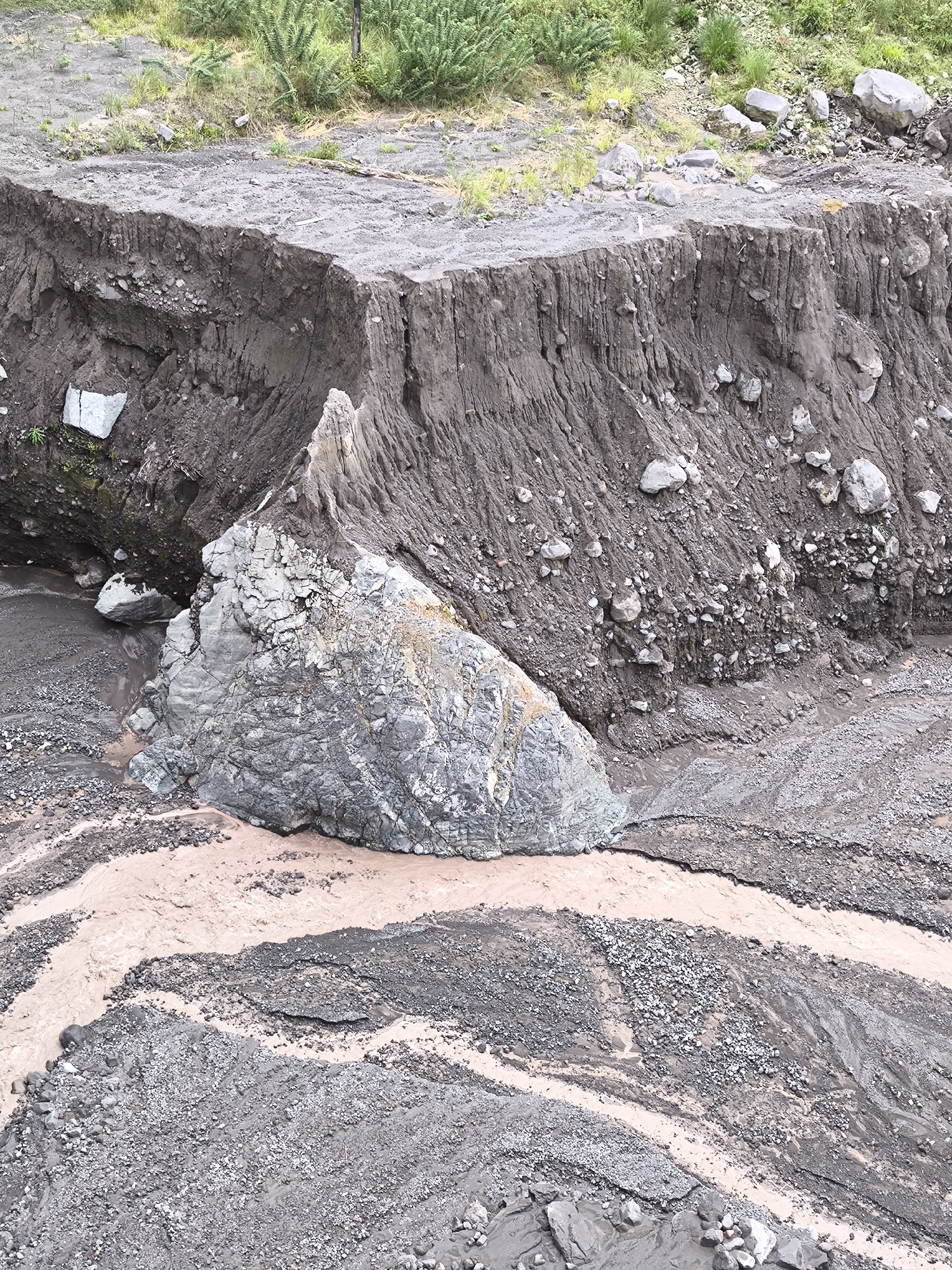 This image depicts a dramatic landscape in Supiturang, Indonesia, characterized by a significant landslide and a flowing river. The scene is dominated by steep, eroded earth banks, revealing layers of soil and rock. A massive, gray boulder, fractured and weathered, sits prominently in the foreground, partially submerged in the reddish-brown water of a river that carves through the debris. The riverbed itself is a mix of fine sediment and scattered stones, with channels of water flowing between the deposited material. Vegetation, sparse and hardy, can be seen on the upper slopes, indicating a rugged, natural environment. The overall impression is one of raw, untamed nature, where geological forces have shaped the land in a powerful and visible manner. There are no people visible in the image.