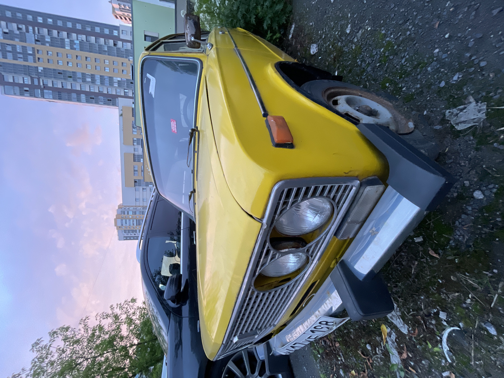 A yellow vintage car is parked on a gravel surface next to a dark SUV. The yellow car has a chrome grille, two round headlights, and a license plate that reads "W7VE 198". The car's front bumper is silver and black. Behind the cars, a tall, modern apartment building is visible against a cloudy sky with pink and blue hues. There are also trees and other buildings in the background. The ground around the cars is covered in gravel, dirt, and some scattered debris. The lighting suggests it is either dawn or dusk.