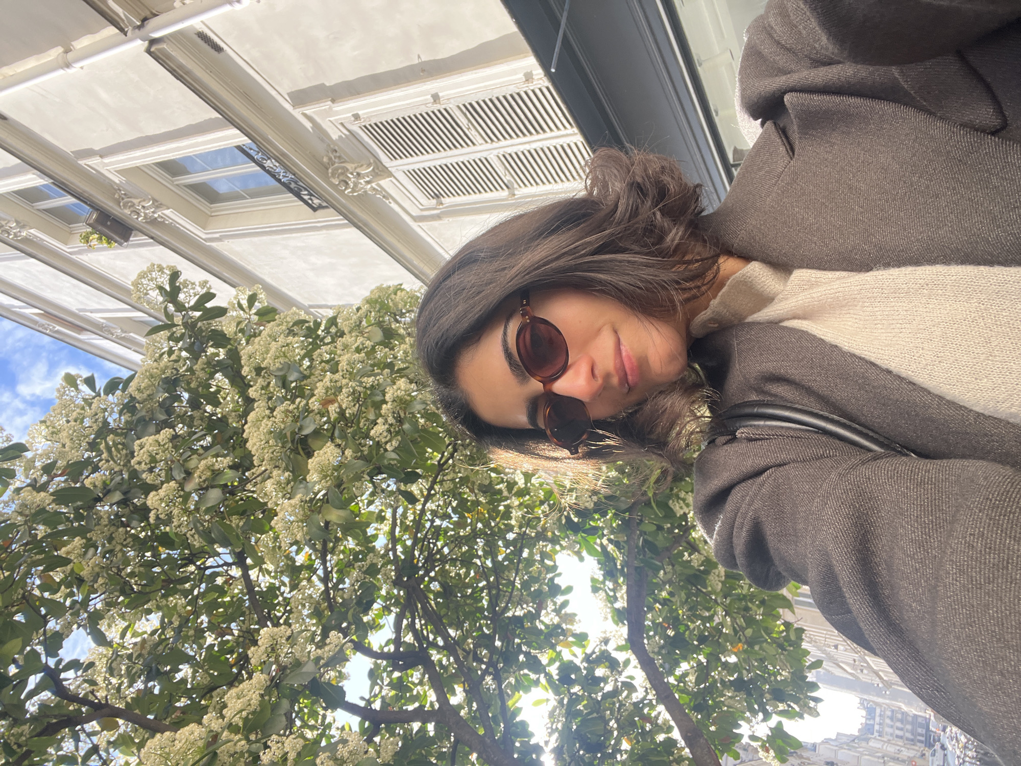 A woman is depicted taking a selfie outdoors, holding the camera from a low angle and looking slightly upwards. She has dark hair, is wearing brown-framed sunglasses, a dark brown or charcoal-grey textured coat, and a light beige or off-white knitted sweater. A dark bag strap is visible over her shoulder.
The scene is set in what appears to be a street or park area in Paris, France, suggested by the architectural style. To the woman's left and center, a vibrant tree with abundant green leaves and clusters of small white blossoms dominates the frame, illuminated by bright sunlight filtering through its branches. Patches of clear blue sky with some clouds are visible through the foliage.
In the background, positioned directly above the woman in a correctly oriented view, are the lower facades of elegant, light-colored buildings. These structures feature ornate cornices, decorative architectural elements, and distinct ventilation grates, characteristic of Parisian Haussmannian architecture. Some windows or skylights on the buildings reflect the blue sky.
The photo captures a bright, sunny daytime scene, likely during springtime given the blooming trees. The woman's expression is calm, with a hint of a smile, enjoying the pleasant outdoor environment. No visible text is present in the image.