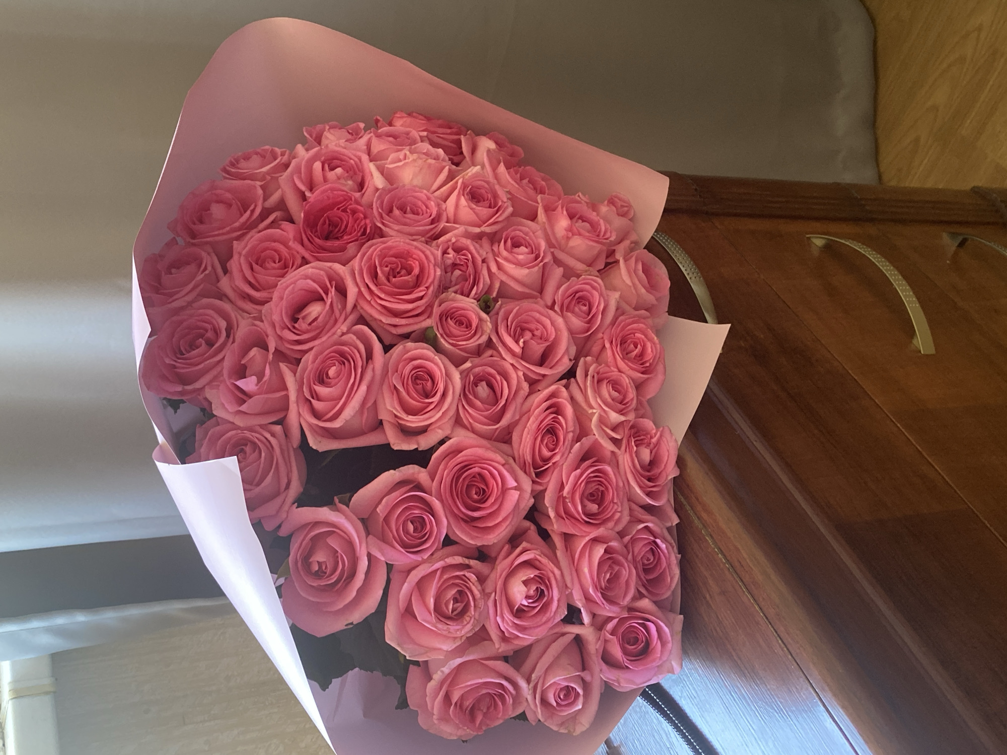 A large bouquet of pink roses, wrapped in pink paper, is positioned in front of a wooden cabinet with metal handles. The location is potentially Sochi, Russia. The environment is indoors.