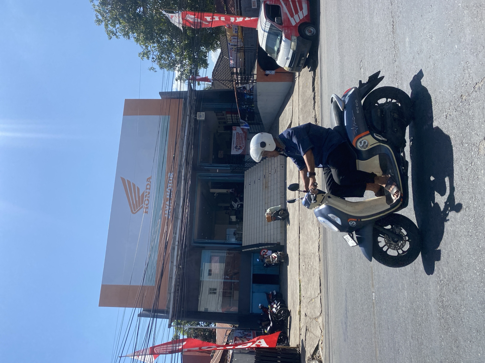 In Bantaeng, Indonesia, on a bright, sunny day, a man wearing a blue shirt, black pants, and a white helmet rides a dark gray and beige scooter down a paved road. The scooter, identified by the "SCOO" label on its rear, is casting a shadow on the road. To the left of the road is a Honda motorcycle dealership named "JAYA MOTOR". The dealership features a large sign with the Honda logo and lettering. In front of the dealership, several motorcycles are parked on the sidewalk. A red and white van, also with Honda branding, is parked on the right side of the road. Red flags are visible on poles in front of the dealership and also to the right of the dealership. The environment appears to be a commercial area with buildings and trees visible in the background.
