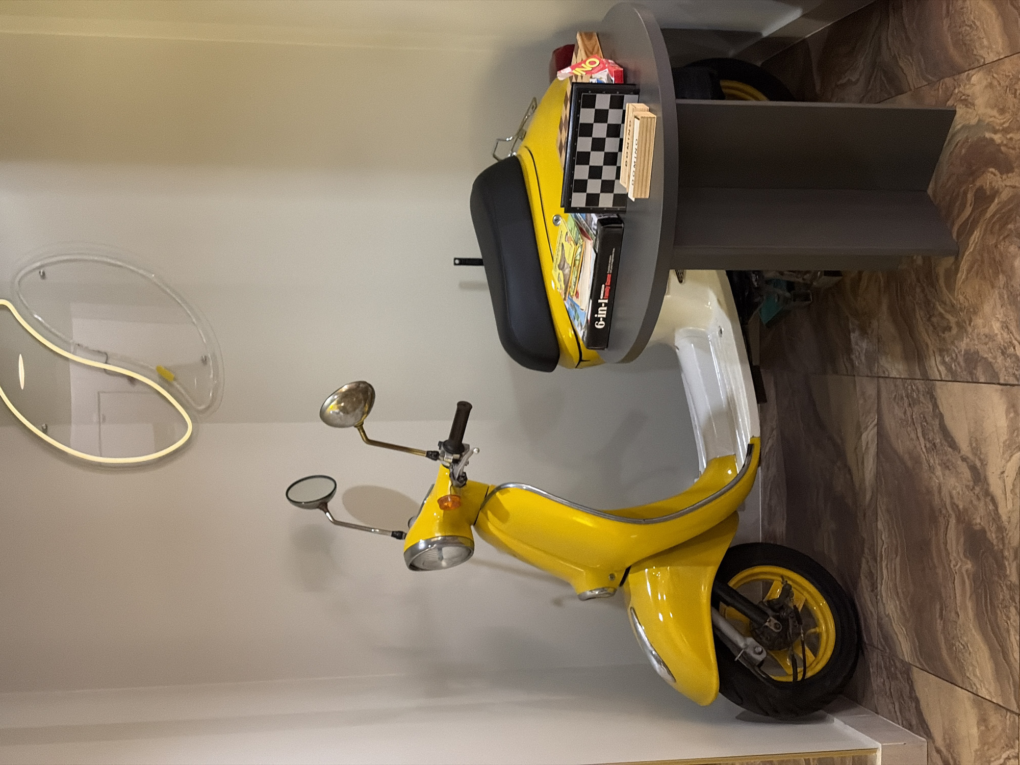 The image shows a room with a yellow Vespa-style scooter attached to a gray side table, seemingly as a decorative furniture piece. The scooter is positioned upright with the rear tire touching the floor and the front facing upward. Games and other items are placed on top of the seat area. The room has a light-colored wall, an oval-shaped decorative lighting fixture and brown tile on another wall. The location is Minsk, Belarus. There are no people visible in the image.