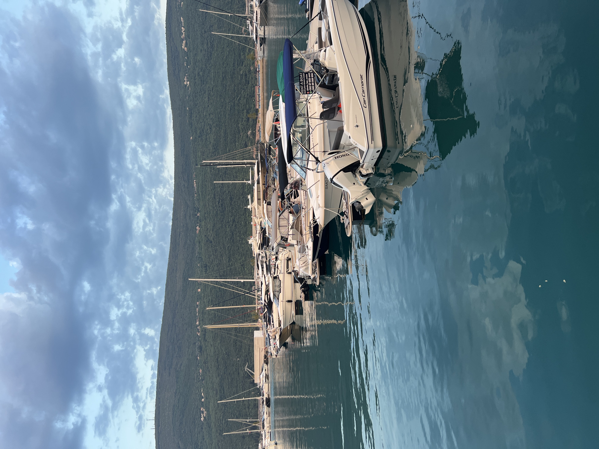 A vertical shot captures a serene marina scene in Đenovići, Montenegro, with a line of boats docked along the water. The boats, varying in size and style, are mostly white with dark trim, and many are equipped with outboard motors. One prominent boat in the foreground has "Cap Camarat" written on its side, and another visible engine bears the "Honda" logo. A sign on one of the boats advertises "RENT A BOAT" with a phone number and website, indicating a commercial operation.
The water is calm and reflects the sky, the boats, and the lush green mountains that rise steeply behind the marina. The mountains are densely covered in trees, and small houses are scattered along their slopes, hinting at a developed but still natural landscape. The sky is partly cloudy, with a mix of bright white cumulus clouds against a pale blue background, suggesting a pleasant, possibly late afternoon, time of day. The overall impression is one of tranquility and a typical summer day at a coastal resort. No people are clearly visible in the image, and the focus is on the boats and the surrounding environment.