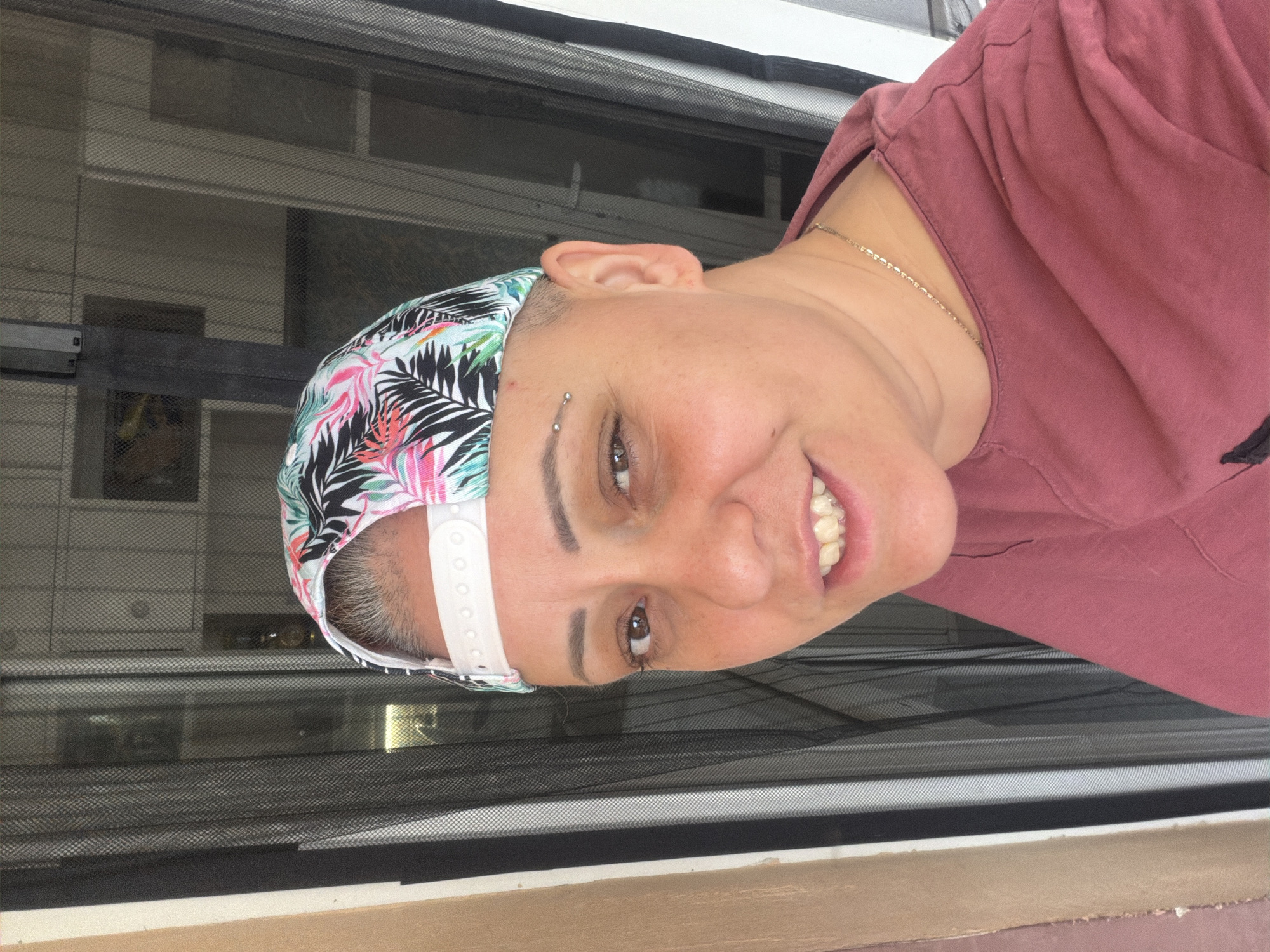 This rotated selfie captures a smiling individual in a close-up shot, presumably taken during the daytime. The person has a very short, buzz-cut hairstyle and is wearing a multi-colored baseball cap, adorned with a vibrant tropical leaf pattern in pink, green, and black on a white base. The cap is worn backwards, with its adjustable white strap clearly visible.
The subject is also wearing a reddish-brown or mauve t-shirt and a thin gold chain necklace. A notable detail is a piercing on their left eyebrow (viewer's right). They are smiling broadly, revealing their teeth, which appear to have clear dental aligners or retainers. This expression conveys a sense of happiness and cheerfulness.
The background primarily features a dark mesh screen, suggesting a window or a screened outdoor area like a porch or balcony. Beyond the screen, blurred shapes hint at other structures or an outdoor scene. There is no visible text in the image. The setting is consistent with a location such as Varna, Bulgaria.