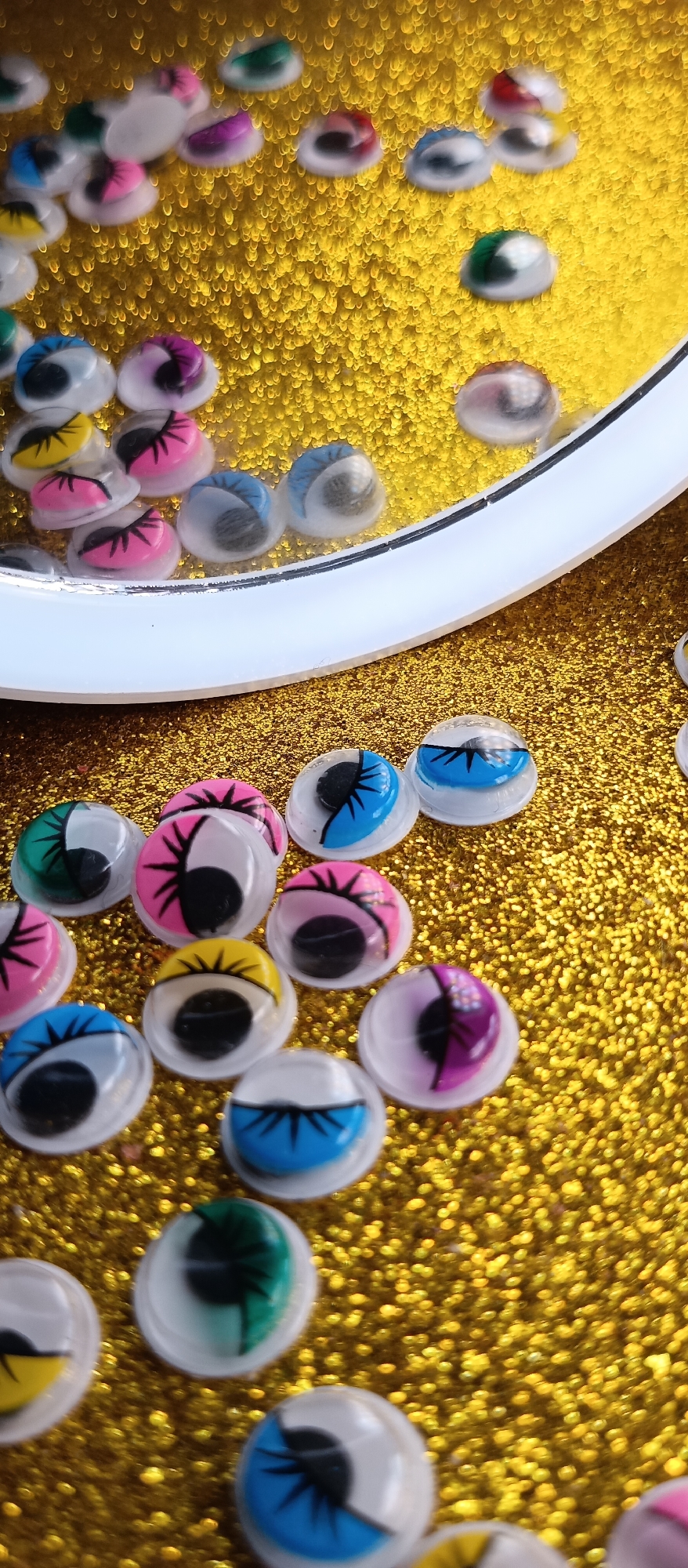 The image presents a close-up, high-angle view of numerous googly eyes scattered across a dazzling golden glitter background. A significant portion of the frame is dominated by a brilliant, sparkling gold surface made up of fine, reflective glitter particles, creating a bokeh effect in some areas.
The primary subjects are the googly eyes themselves, which come in a variety of vibrant colors. Many of the eyes feature colored eyelids or irises in hues such as pink, blue, yellow, green, and purple, often combined with black pupils and distinct black eyelash or spike designs. Some eyes exhibit a half-moon design where one half is colored and the other is a plain white or clear dome. The eyes are small, round, and have a clear, domed plastic casing.
A prominent white, curved object, resembling the rim or edge of a shallow container or shaker, cuts across the middle of the image. Inside this curved enclosure, several googly eyes are visible, appearing slightly out of focus and seemingly suspended, suggesting they are either floating in liquid or part of a shaker mechanism. Outside and in the foreground of this rim, a larger collection of googly eyes is arranged directly on the glittery surface, appearing sharper and more detailed.
There is no discernible activity or event taking place; the image is a still life composition. The setting appears to be indoors, possibly on a craft table. The lighting is bright, emphasizing the reflective qualities of both the glitter and the smooth surfaces of the googly eyes. No text is visible in the media file. The overall impression is one of playfulness and vibrancy, typical of crafting materials or decorative elements.