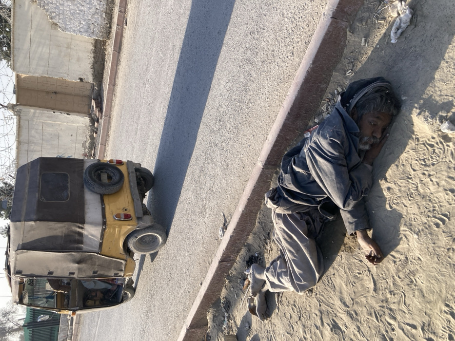 An overhead view captures a scene on a sunny day in Quetta City Tehsil, Pakistan. In the foreground, a person lies prone on the sandy ground, parallel to a reddish-brown curb. The individual is dressed in light grey trousers and a dark blue or black long-sleeved garment, with a dark cloth or turban wrapped around their head. Their face is visible, turned slightly towards the viewer, with eyes partially open. They wear light-colored open-toed sandals or chappals on their feet.
Immediately behind the curb is an asphalt road. On the road, slightly to the left of the frame's center, a yellow and dark-colored three-wheeled auto-rickshaw is positioned with its rear facing the viewer. The vehicle has a canvas top, a visible spare tire mounted on the back, and appears to have a single occupant in the driver's seat. Long shadows are cast across the road to the right.
In the background, beyond the road, a light-colored concrete wall topped with barbed wire runs horizontally. A reddish-brown gate is visible in the wall. Sparse foliage or trees are visible further in the background to the left.