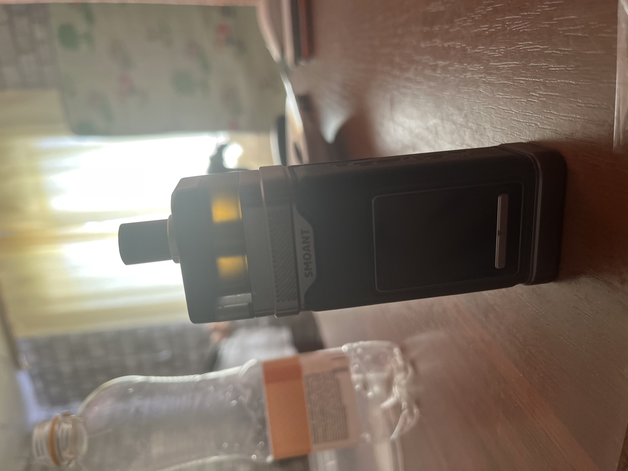A black vape device with "SMOANT" branding sits on a wooden table. To the left of the vape is a clear plastic bottle with a white label containing orange text and several small air bubbles inside. In the background, a window is covered by light yellow curtains, and a patterned chair is visible to the right. The scene appears to be indoors, potentially in a residential setting in Baykalsk, Russia.