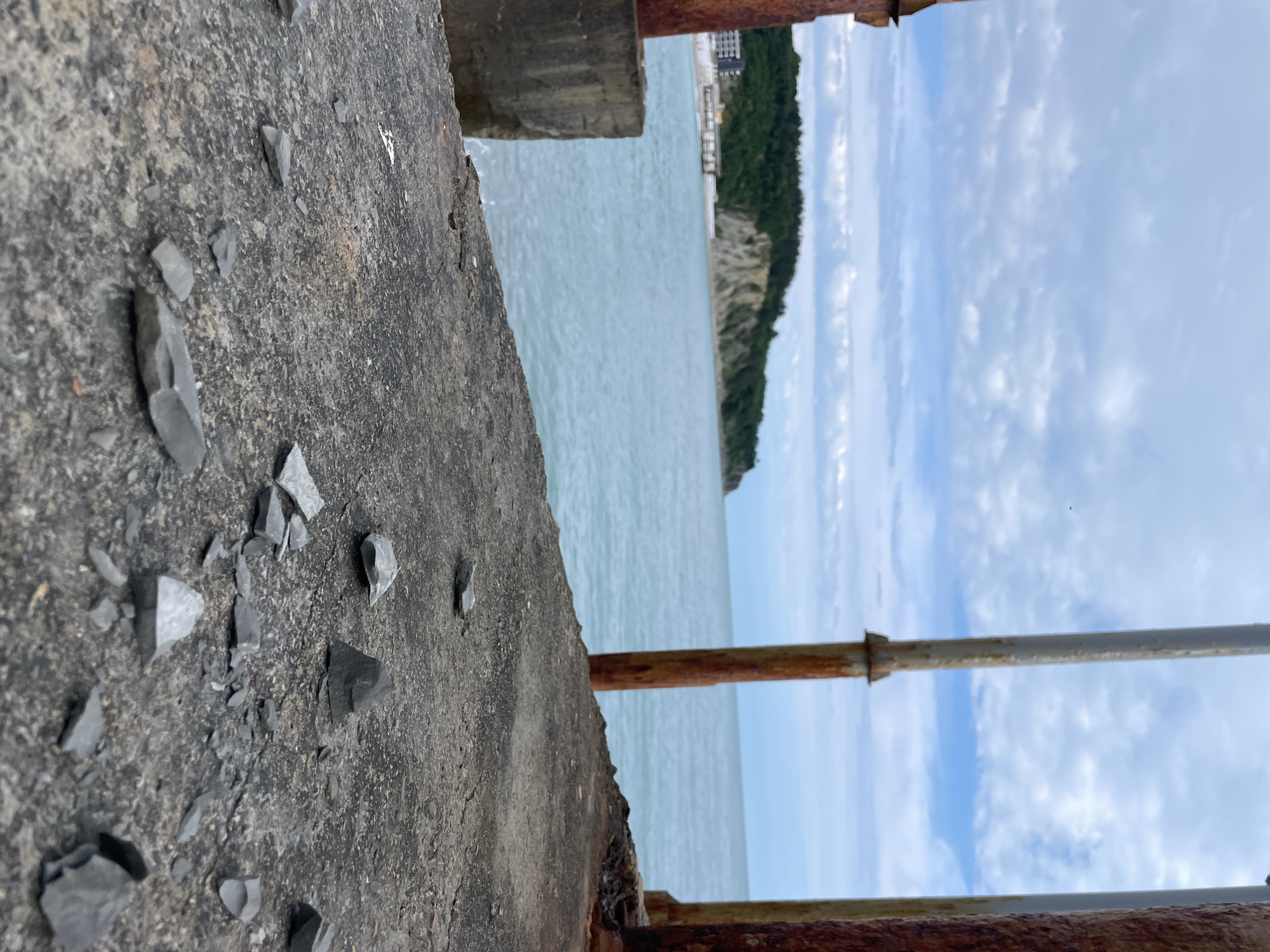 The image depicts a close-up view of a concrete ledge or wall with scattered dark, angular fragments. Rusted metal railings are present in the foreground. Beyond the ledge, the scene opens to a body of water, possibly the sea, with land visible in the distance. The sky is cloudy with patches of blue.
While the image's location is stated to be Olginka, Russia, the provided visual information alone does not confirm this location. Additional contextual clues would be needed to verify this claim.