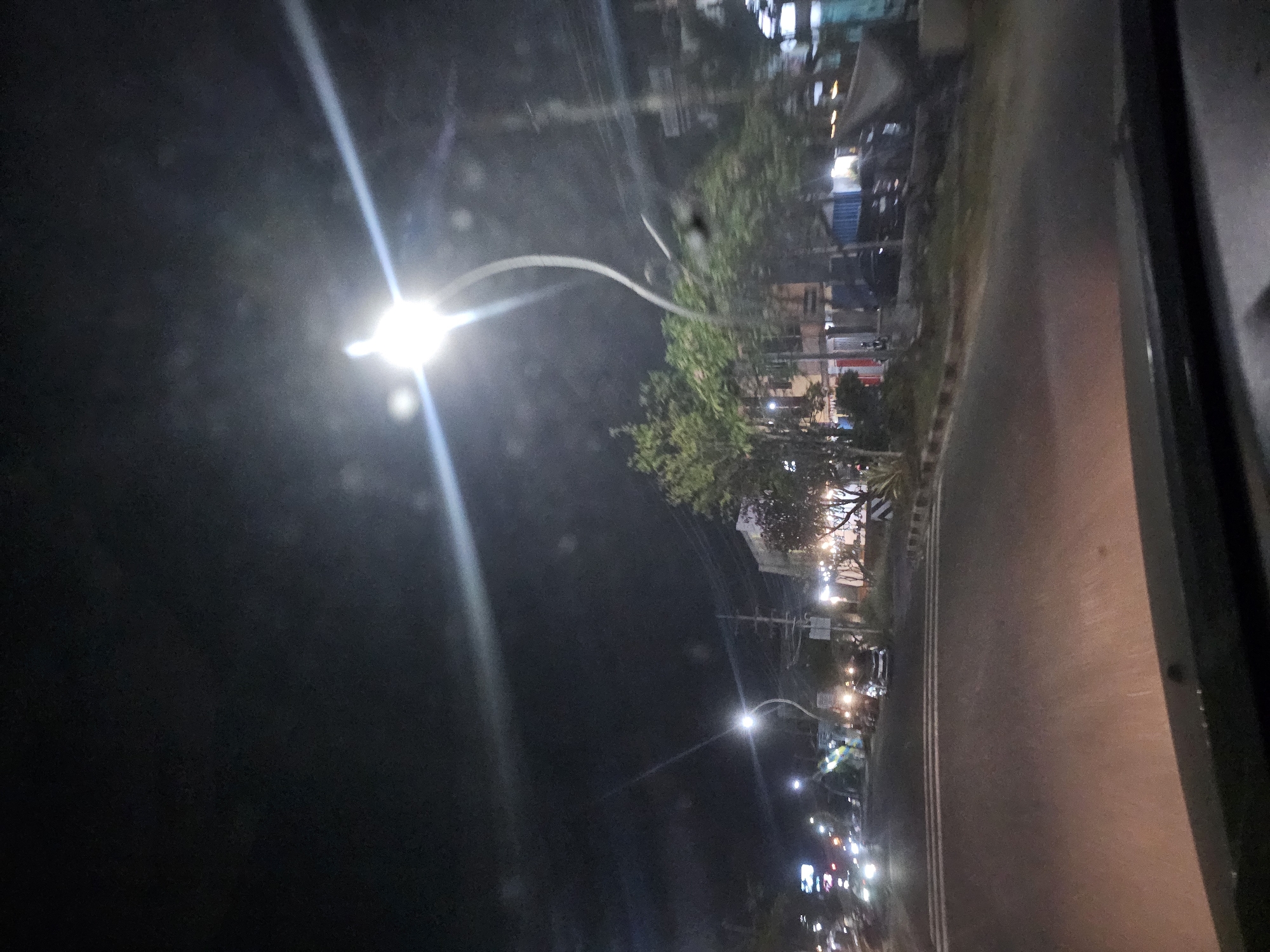 The image depicts a nighttime scene on a street in Sukomulyo, Indonesia. The perspective is from within a vehicle, looking forward. A streetlamp with a distinctive curved pole illuminates the road and the surrounding area. On the left side of the road, there are buildings with illuminated signs, and several cars are visible, some with their headlights on. The right side of the road features trees and more buildings, some with lights indicating businesses or residences. Power lines are visible overhead, supported by utility poles. The road surface appears to be asphalt, with white lane markings. The overall atmosphere suggests a typical urban environment at night.