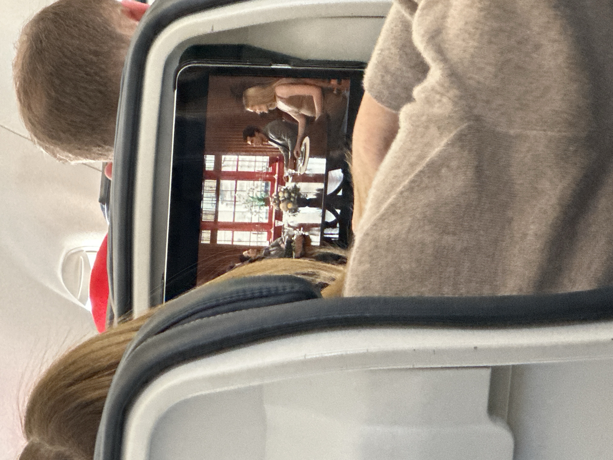 The image shows a partially obstructed view of an airplane passenger using a personal device to watch a video. The screen displays a scene of a man and a woman at a dining table in front of a window. The scene appears indoors and does not provide any indication of being located in Sant Josep de sa Talaia, Spain. There is no specific evidence within the image to confirm the location in Sant Josep de sa Talaia, Spain. The passenger is seated behind another person, whose hair is visible near the bottom left of the frame. The head of another passenger can be seen to the left of the screen. The arm and shoulder of the person sitting to the right is visible.