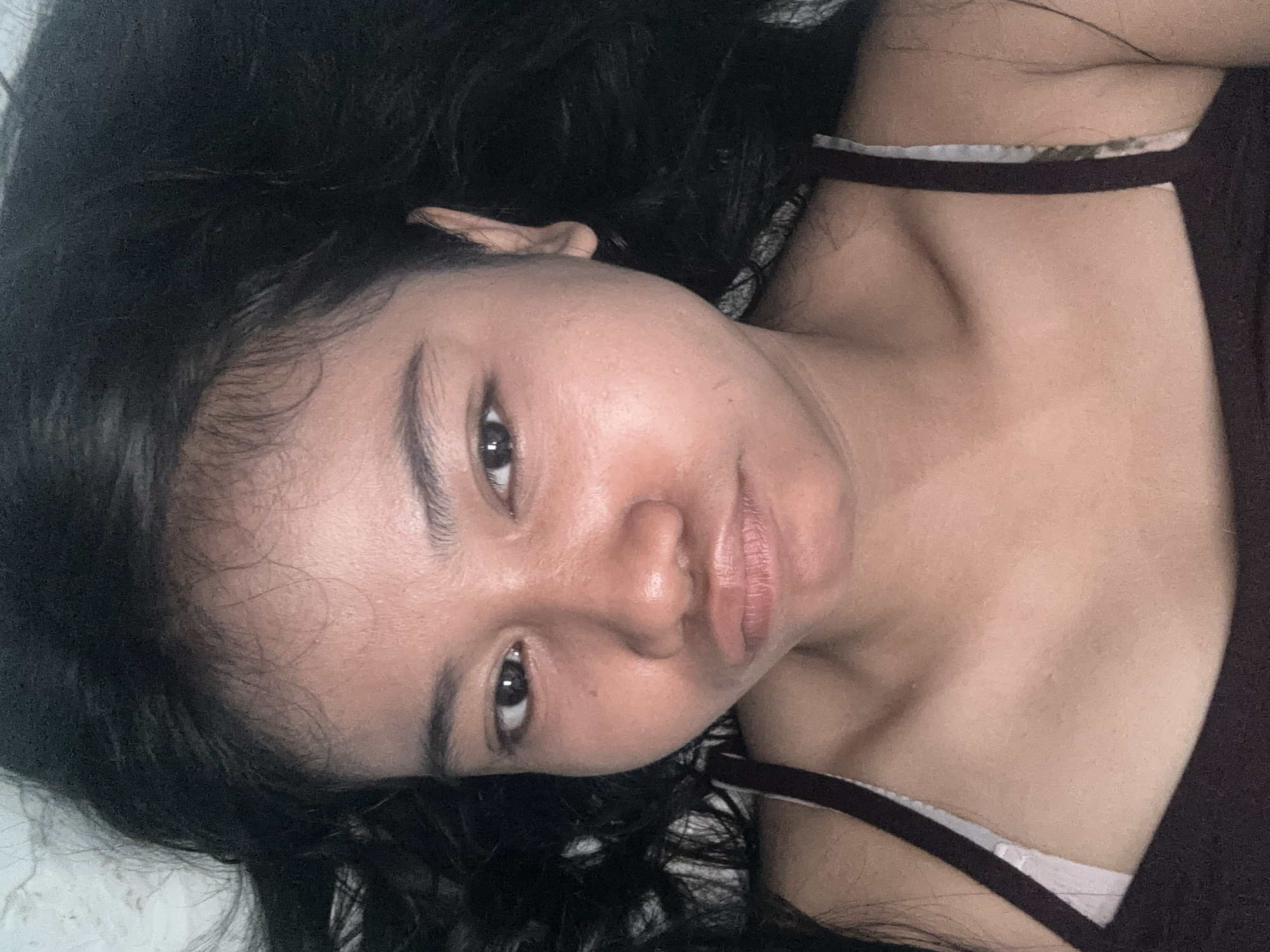 The image is a close-up, overhead selfie of a young woman with dark hair, lying down. She is wearing a dark tank top with thin white straps visible on the shoulder. Her facial expression is a pout. The background is indistinct, suggesting an indoor setting, possibly a bed. The lighting is soft and appears to be from overhead. There are no discernible city landmarks or specific environmental details that would allow for precise location identification within North Jakarta, Indonesia. The focus is entirely on the subject's face and upper torso.