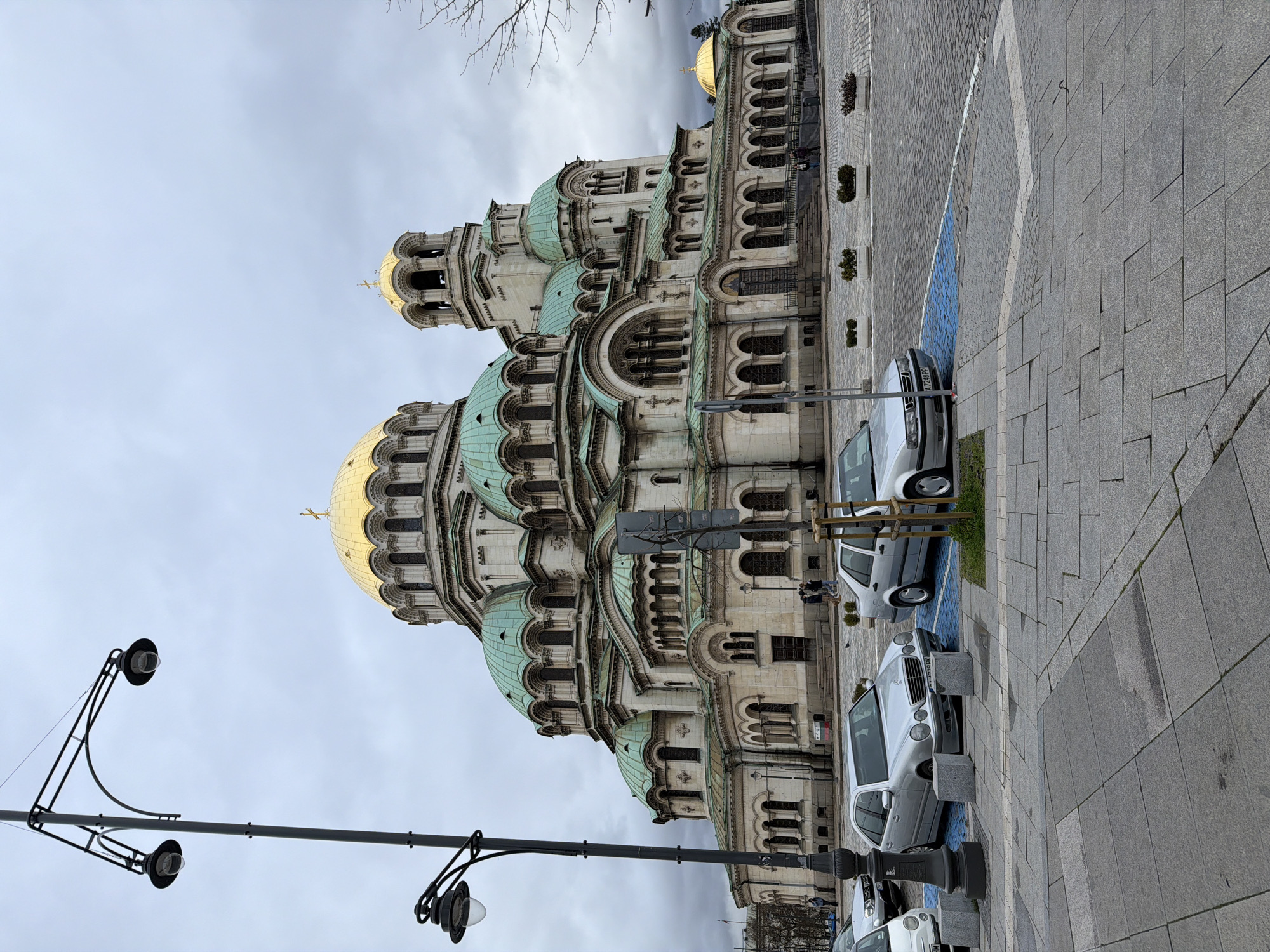 The image depicts the Alexander Nevsky Cathedral in Sofia, Bulgaria. The cathedral, a large Eastern Orthodox church, dominates the left side of the image with its gold-covered domes and green rooftops. A row of parked cars, predominantly silver, lines the right side of the frame along a paved walkway. A black street lamp is located on the left side, close to the foreground, while the sky is overcast.