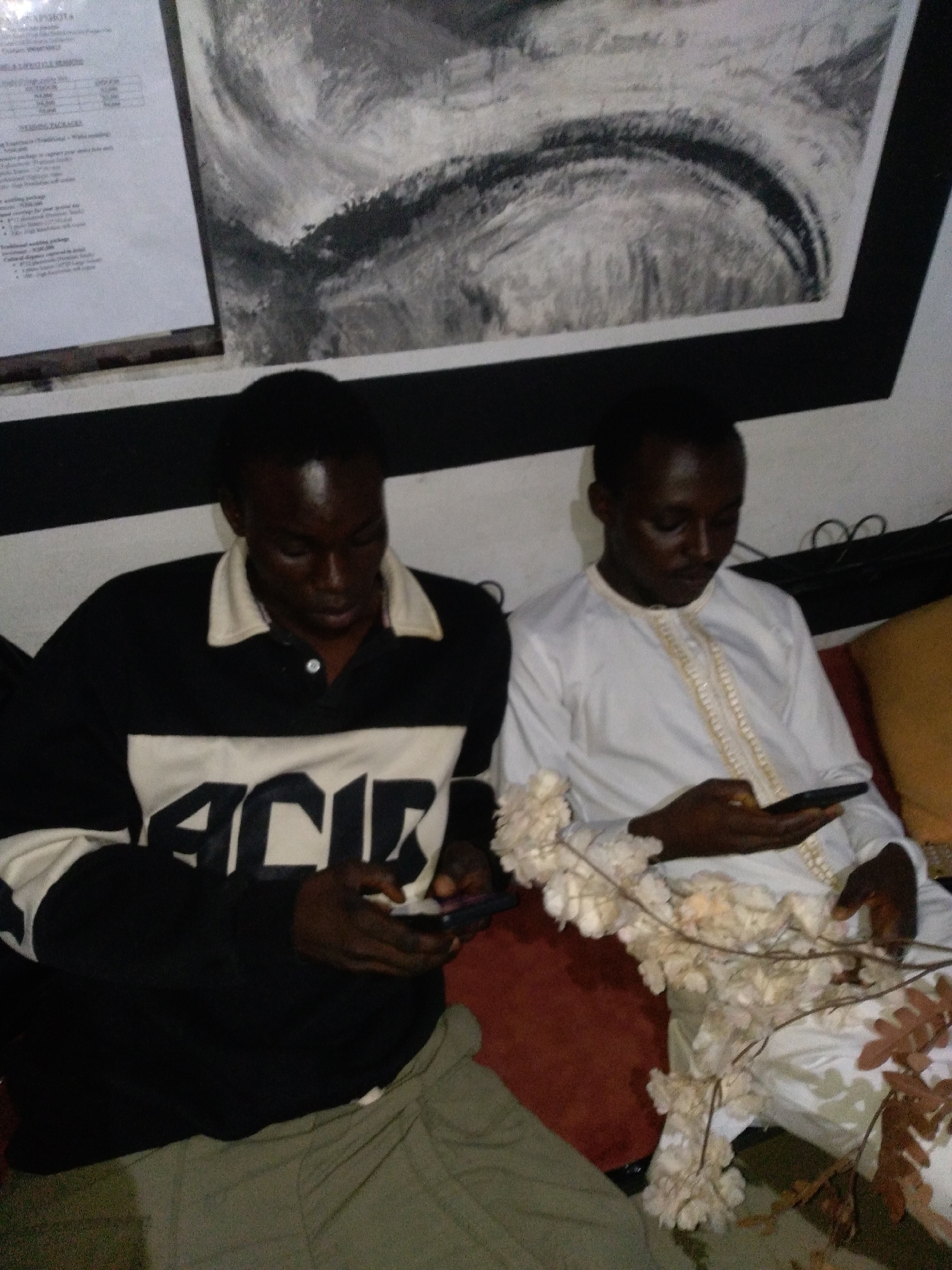 An indoor scene features two men of dark complexion seated side-by-side on a reddish-brown upholstered surface, each engaged with a smartphone, in the city of Unknown, Unknown. The man on the left wears a black long-sleeved polo shirt with a white collar and a broad white horizontal stripe across the chest, which displays dark block letters, likely "ACID." He is wearing olive-green pants and holds his dark-colored smartphone with both hands, looking downwards. The man on the right wears a white traditional garment, possibly a thobe, adorned with intricate gold embroidery along the front placket. He holds a dark-colored smartphone in his right hand, also looking downwards. A long strand of white, textured, possibly artificial flowers or dried plant material is draped across his lap and extends towards the left. A branch with brown, dried leaves is visible in the lower right foreground. The background consists of a wall with a prominent dark horizontal stripe. Above this stripe, on the left, a white framed document with black text, possibly a menu or price list, is partially visible. To its right, a large, framed abstract painting in black, white, and gray tones, featuring curved brushstrokes, is displayed. The overall lighting is dim.