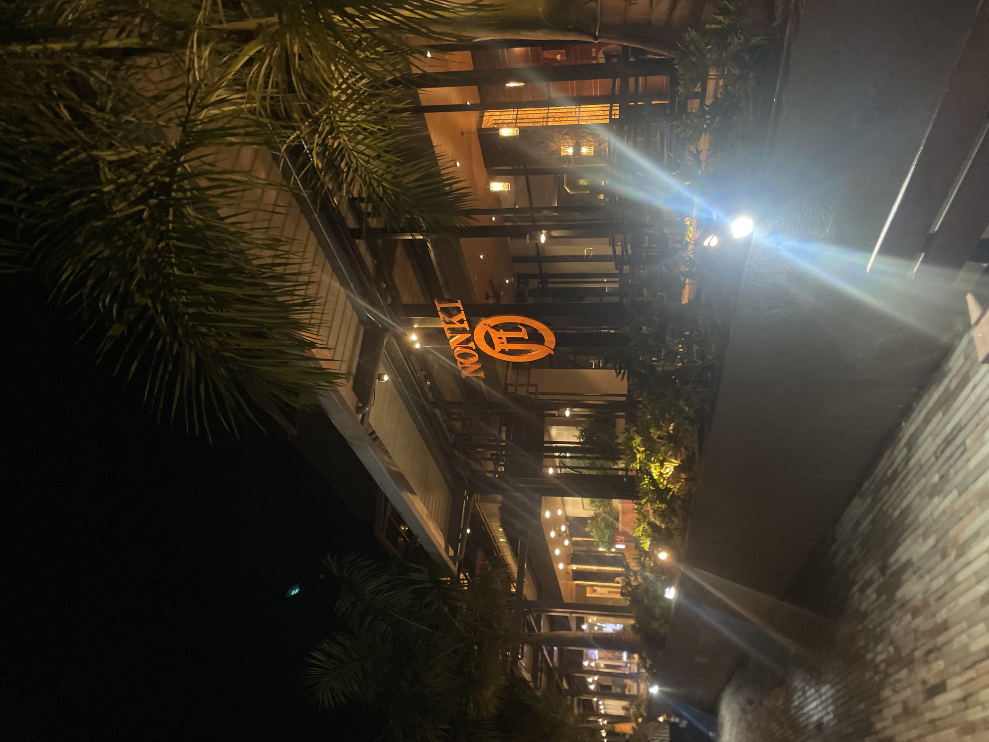 At night, a view of a building with the brightly lit orange sign "NONKI" is visible through palm trees. The building has multiple floors with visible interior lighting. The background is dark, and the scene includes a brick structure to the right of the frame. The location is in the city of Unknown, Unknown.