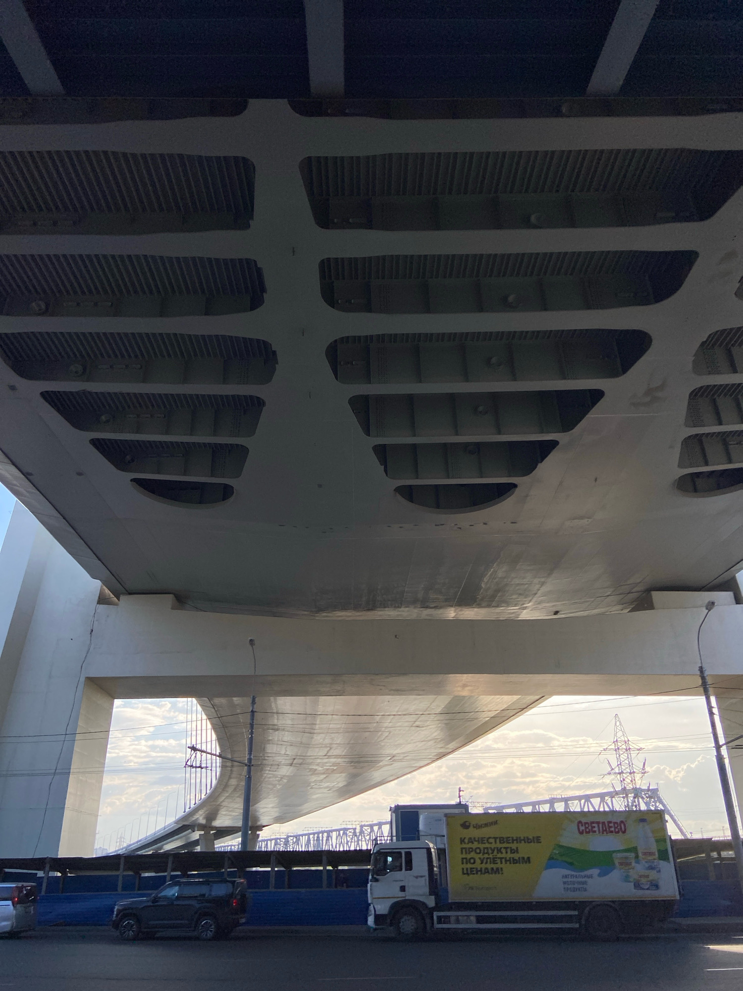 This media file depicts an underpass scene beneath a large concrete bridge structure. The underside of the bridge is characterized by intricate ribbed metal supports and a smooth, curved concrete surface with several large rectangular and oval cutouts.
In the foreground, a dark-colored SUV is parked on the street. To the right, a white truck with a yellow advertising banner is visible. The banner features text in Russian, including "СВЕТАЕВО" (Svetaevo), "КАЧЕСТВЕННЫЕ ПРОДУКТЫ ПО УЛЁТНЫМ ЦЕНАМ!" (Quality Products at Awesome Prices!), and "НАТУРАЛЬНЫЕ МОЛОЧНЫЕ ПРОДУКТЫ" (Natural Dairy Products).
Beyond the truck, a blue barrier runs along the street, and further in the background, the curved structure of another bridge or elevated roadway is visible, along with a large electrical pylon. The sky is partly cloudy, suggesting it is daytime, likely late afternoon due to the warm lighting and long shadows. The overall scene conveys a sense of urban infrastructure and commercial activity, set against a backdrop of a large bridge. The location context is Novosibirsk, Russia.