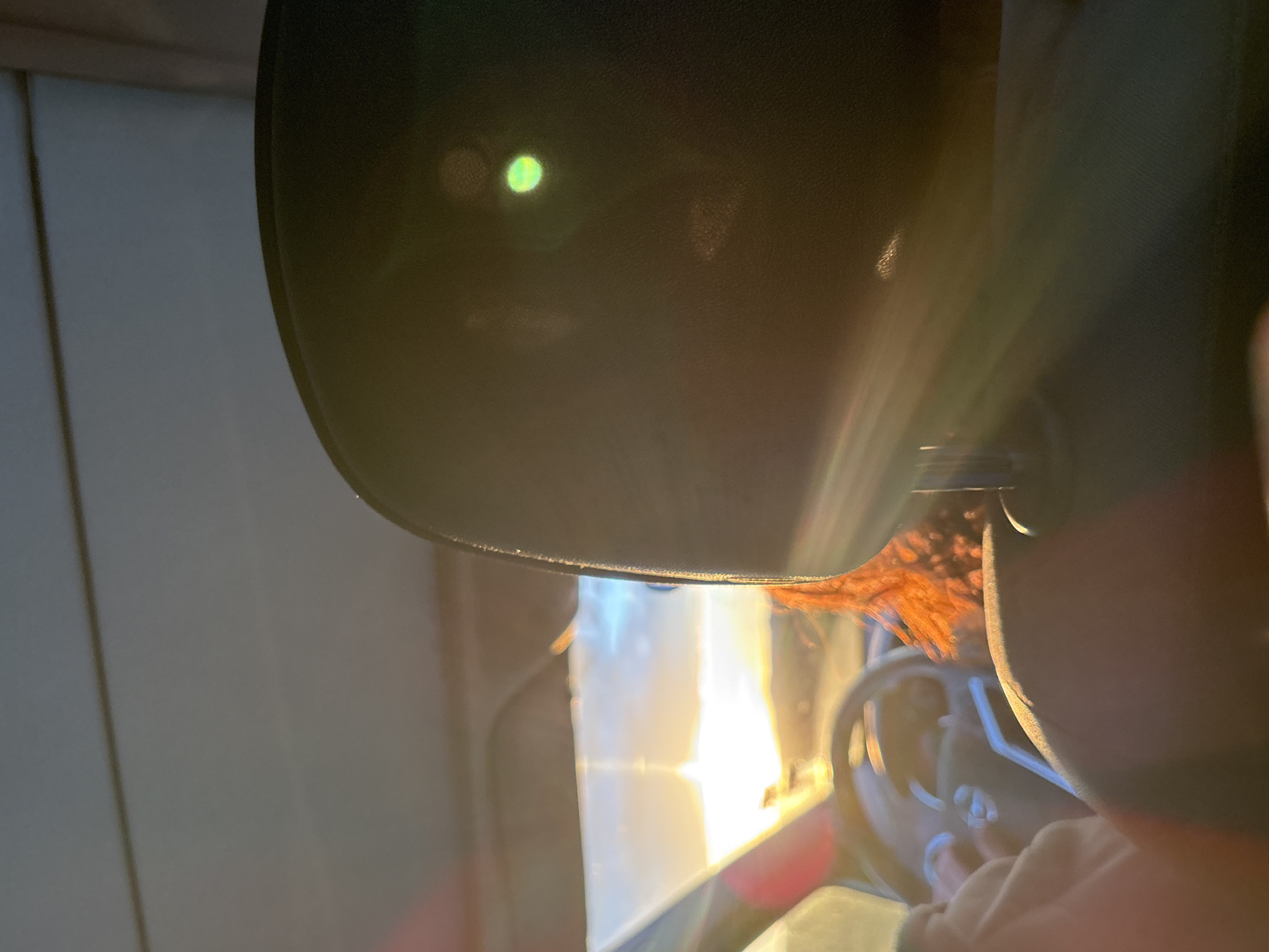 The image appears to be taken from inside a vehicle. A dark, possibly black, visor is partially obscuring the view. Through the windshield, a bright light, possibly sunlight, is visible. The steering wheel and hands are visible, indicating someone is driving. There is reddish-orange hair near the steering wheel. The location is unknown and not identifiable from the available information.
