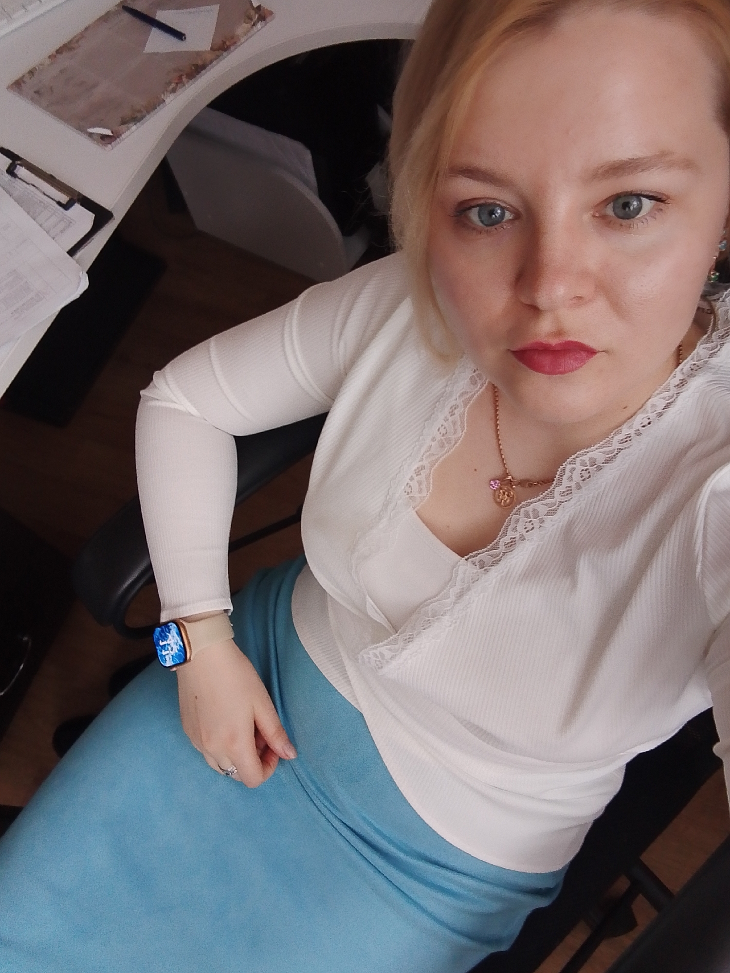 A fair-skinned woman with blonde hair and blue eyes is seated, looking up at the camera. She has red lipstick on. She is wearing a white, long-sleeved, ribbed shirt with lace trim at the neckline, and a light blue skirt. She is wearing a gold necklace. On her left wrist, she wears a smartwatch with a light-colored band. She is sitting in an office chair with black arms. Part of a white desk is visible in the top left corner of the frame, with papers and a pen on it. The location of the photo is in the city of Izhevsk, Russia.