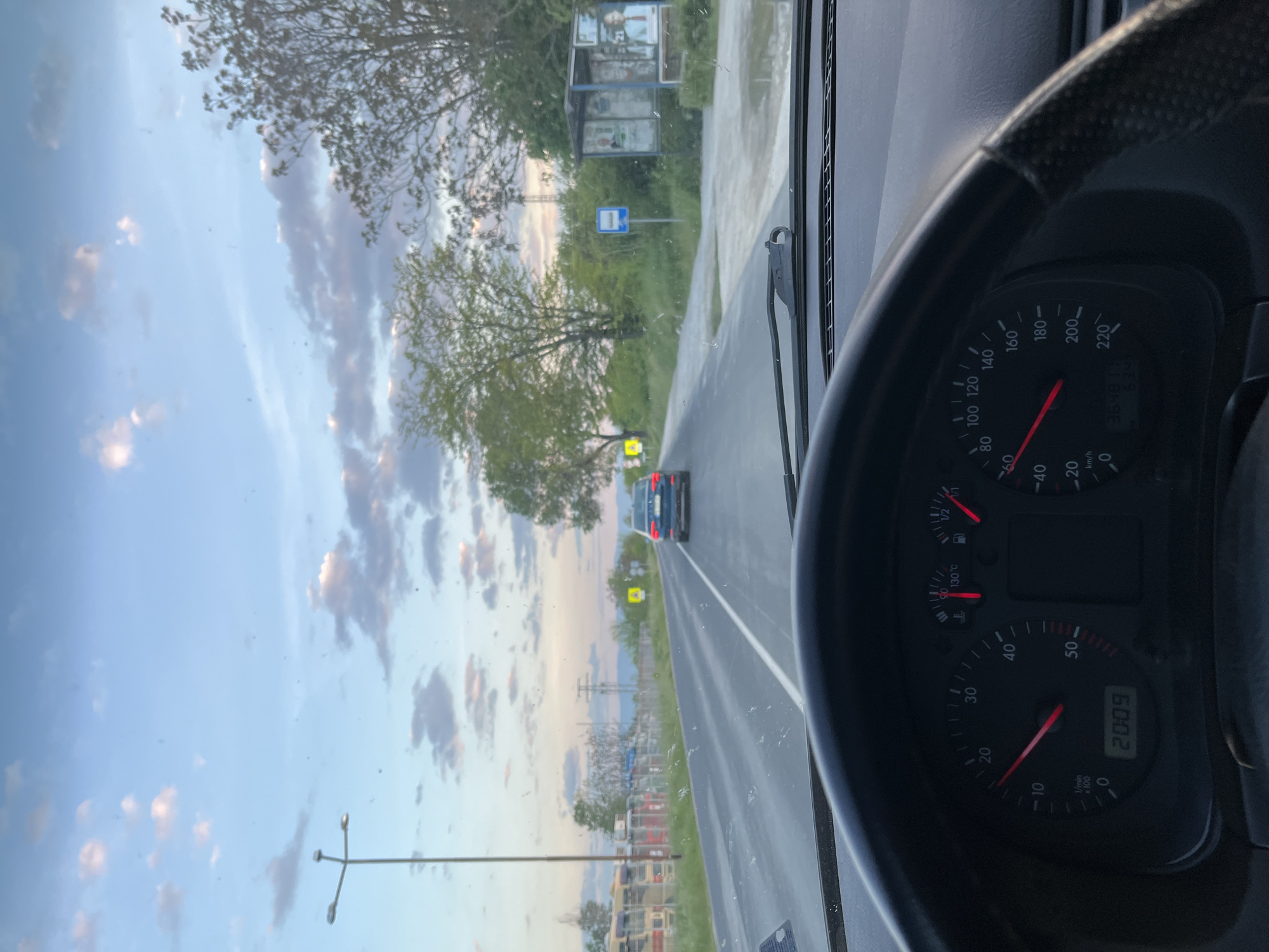 The image, taken from inside a car, depicts a view of a road leading into the distance. The car's dashboard and steering wheel are visible in the foreground, with the speedometer indicating the vehicle is in motion. Through the windshield, the road stretches ahead with a car in the distance and roadside vegetation. A roadside structure, possibly a bus stop, is visible to the left. The sky is filled with clouds. The location may be near Dalgo Pole, Bulgaria, based on potential linguistic clues in the roadside structure.