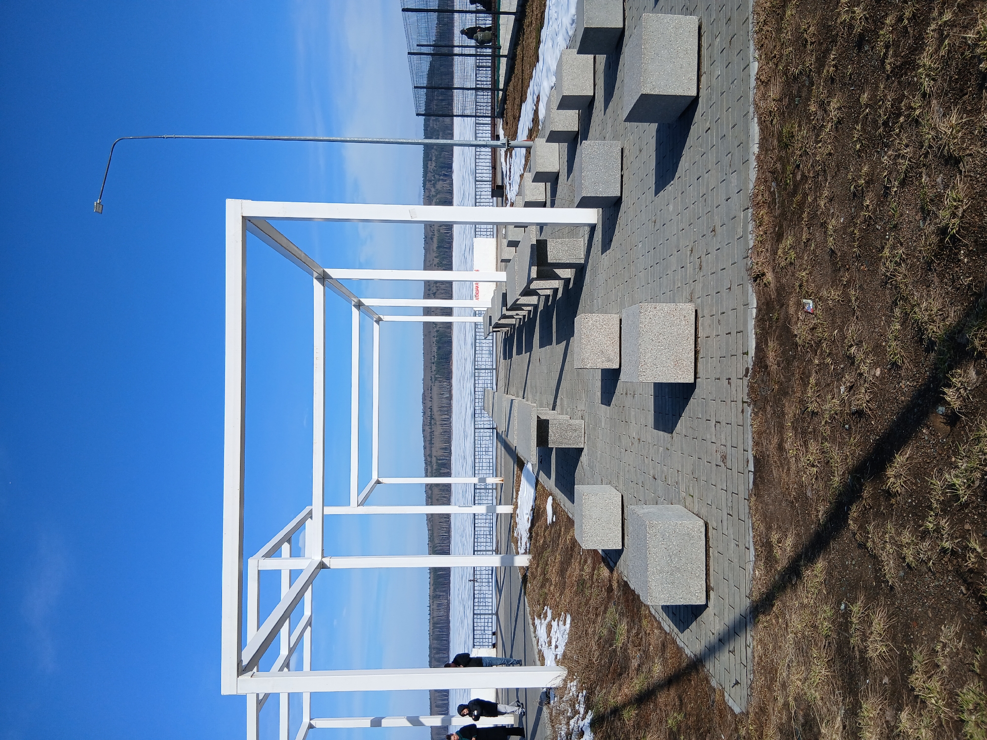The image depicts an outdoor space in an urban setting. A white, cubic metal structure stands prominently, framing a view of a water body, likely a river or a lake, with a partly snow-covered shoreline and a wooded area in the distance. Above, a clear blue sky is visible. To the right of the structure, there is a paved walkway with a series of square stone blocks, presumably for seating. Next to the walkway, there is an area of dry grass and dirt. In the distance, a metal fence runs along the shoreline. Several people can be seen in the lower part of the image, including two individuals near the bottom of the frame. A light post extends upwards on the left side of the image. The location is city of Unknown, Unknown.