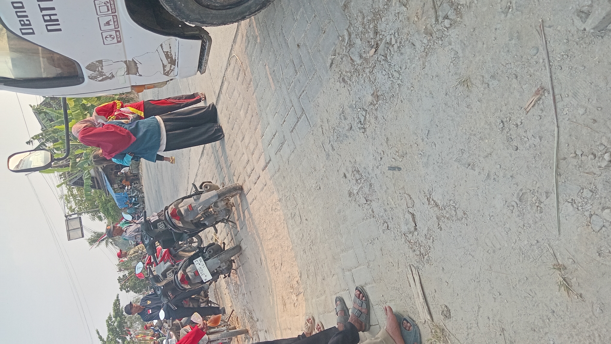 The image shows a street scene in Kedungwaru, Indonesia, with several people and motorcycles. A white truck with text on its side is parked on the right side of the road, partially obscuring a small building and some vegetation in the background. In the foreground, several motorcycles are parked. A group of people are standing near the motorcycles and the truck. One woman is holding a child. The ground is a mix of dirt and paving stones. The sky is bright, suggesting it is daytime.