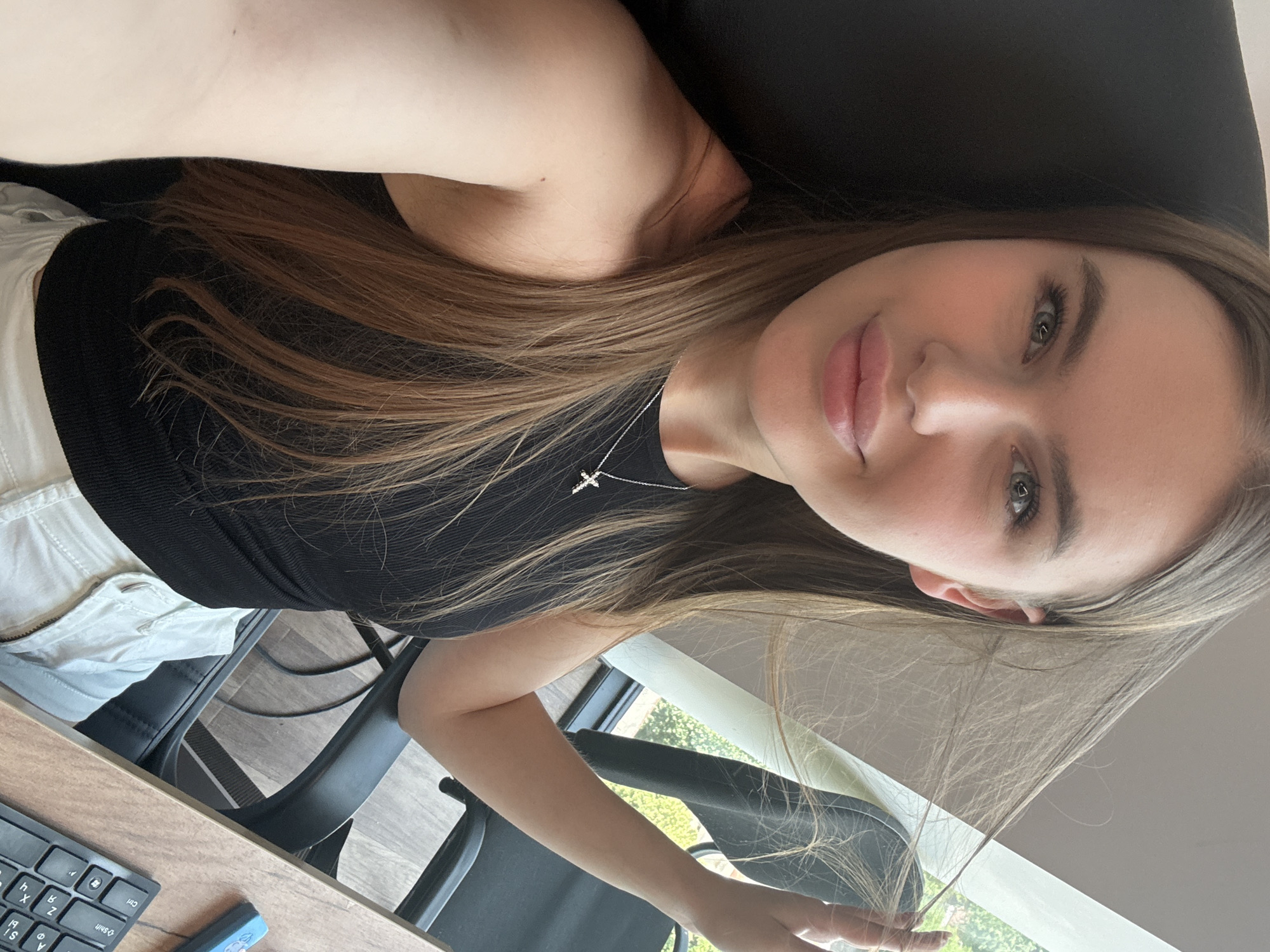 This media file shows a young woman with long, light brown hair taking a selfie. She is wearing a black ribbed tank top and white pants. A silver cross necklace is visible around her neck. Her eyes are light green, and her lips are full and slightly parted. She appears to be in an indoor setting, possibly an office or study, with a desk and keyboard in the foreground and chairs in the background. A window with a view of greenery suggests it is daytime, with natural light illuminating the scene. The overall mood is casual and relaxed. There is no visible text in the image. The location context is Dnipro, Ukraine.
