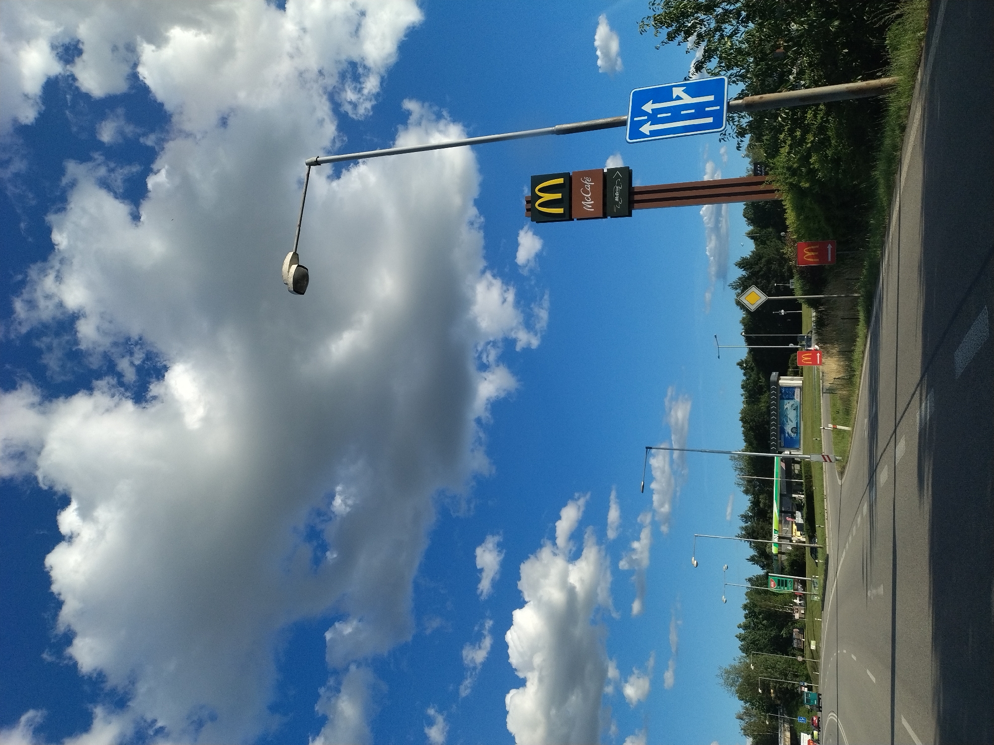 The image shows a sunny day with a bright blue sky and scattered white clouds, possibly in the city of Písek, Czechia. A road curves to the right, with two lanes in each direction marked by white lines. On the left side of the road, there are trees and a gas station with green and white signage. On the right side, a tall pole with a McDonald's sign and a McCafe sign is visible. Adjacent to it, a blue traffic sign with white arrows indicates a lane split, with one lane going straight and the other turning right. There are a few cars visible on the road and signs for McDonald's.