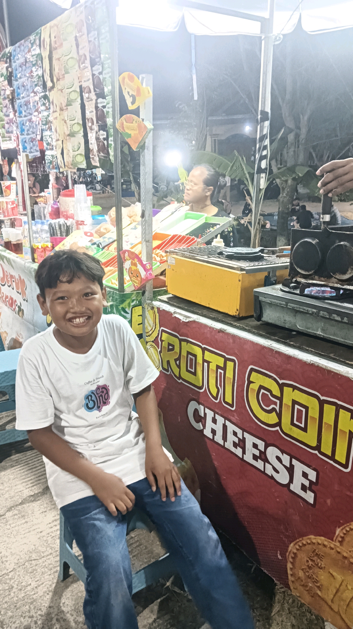 In Newung, Indonesia, a young boy with a wide smile sits on a blue plastic chair in front of a food stall at night. He is wearing a white t-shirt with a graphic and blue jeans. Behind him, the stall displays various packaged snacks and food items. A woman with glasses is visible behind the counter, and a grill with a cooking element is set up. The stall has a large red banner with yellow lettering that reads "ROTI COI CHEESE." The ground is paved, and the overall scene suggests a bustling night market or street food vendor.