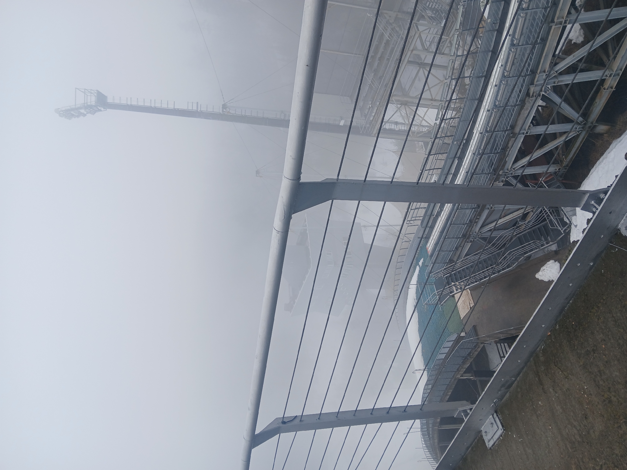 The image shows a metal structure in foggy conditions in Прикамский, Russia. The structure includes a railing, cables, and beams. In the background, a tall metal tower is visible through the fog. The structure appears to be an observation deck or part of a larger industrial complex. There is snow on the ground in a few places.