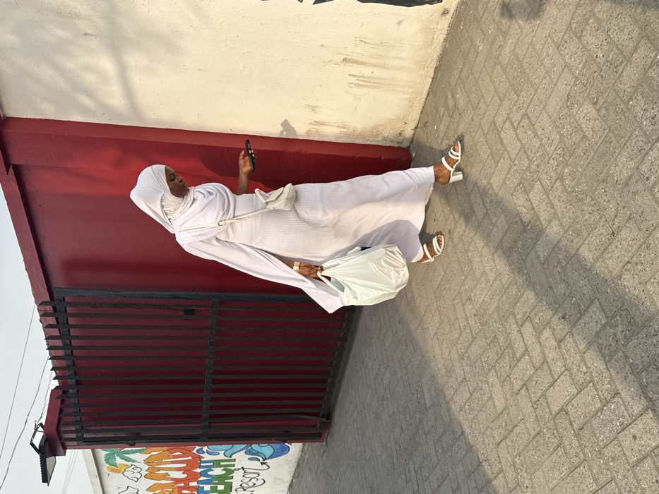 A person dressed entirely in white, including a hijab and sandals, walks along a sidewalk. She holds a phone in one hand and a plastic bag in the other. A small white purse hangs from her shoulder. She is walking alongside a red and white building with a dark metal fence. A colorful mural is partially visible behind the fence. The sidewalk is paved with gray bricks and cast in strong sunlight.