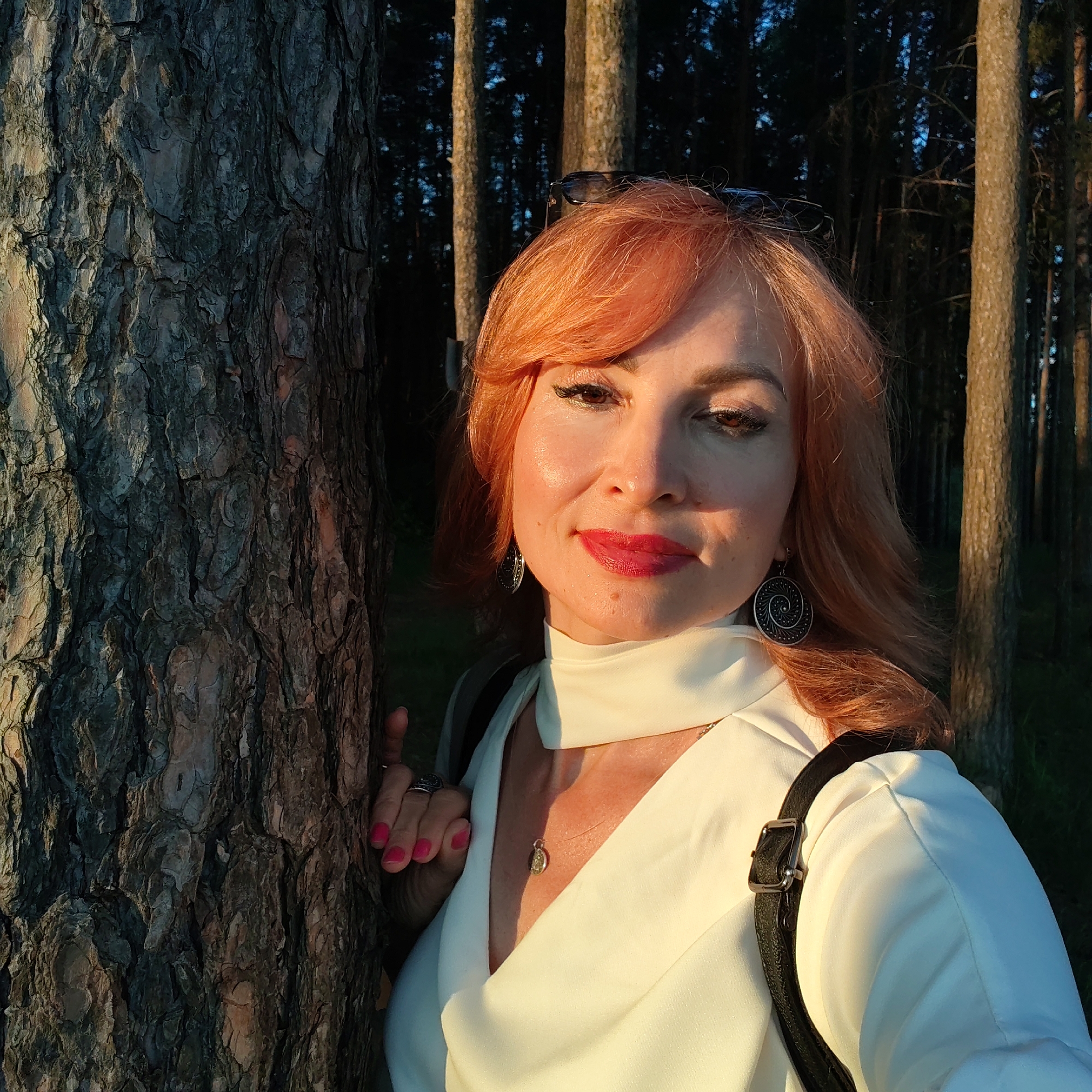 An adult woman with red-orange hair is captured in a close-up selfie-style portrait within a pine forest in Chaikovsky, Russia, during the golden hour. She stands next to a large, textured tree trunk, which frames the left side of the image. Her right hand, visible on the tree bark, has pink nail polish and rings.
The woman exhibits a slight, content smile, accentuated by red lipstick. Her eyes are adorned with dark eyeliner and eyeshadow, and her eyebrows are well-defined. Sunglasses are perched on top of her head. She wears large, circular earrings with a distinctive spiral pattern and a delicate necklace featuring a small pendant. Her attire includes a light-colored (off-white/cream) top with a high, cowl-like collar, and a black bag strap is visible over her right shoulder.
The scene is bathed in warm, directional sunlight, indicating late afternoon. This golden light illuminates the woman's face and the prominent features of the tree bark, creating strong highlights and shadows. The background shows a dense, darker forest composed of tall, slender pine trees, with hints of blue sky peeking through the canopy. The overall atmosphere is natural and serene. No visible text is present in the media file.
