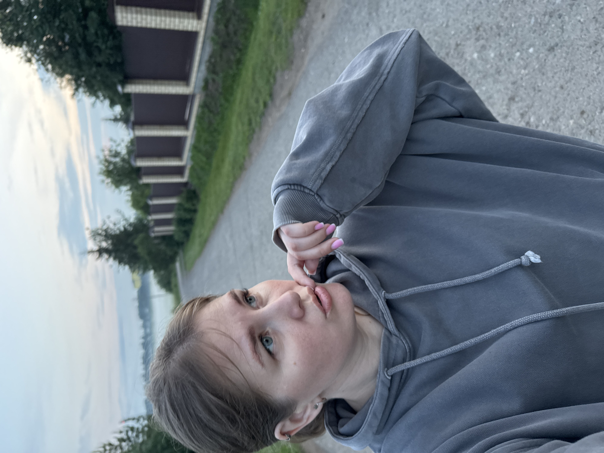 A young woman with light brown hair pulled back stands on a paved road, facing left and looking away from the camera. She is wearing a grey hooded sweatshirt and has her right hand up to her mouth, with her fingers touching her lips. Her fingernails are painted pink. The road appears to be in a rural or suburban setting, with trees and a fence visible in the background. The sky is overcast and appears to be either dawn or dusk. The location is identified as Gorniy, Russia.