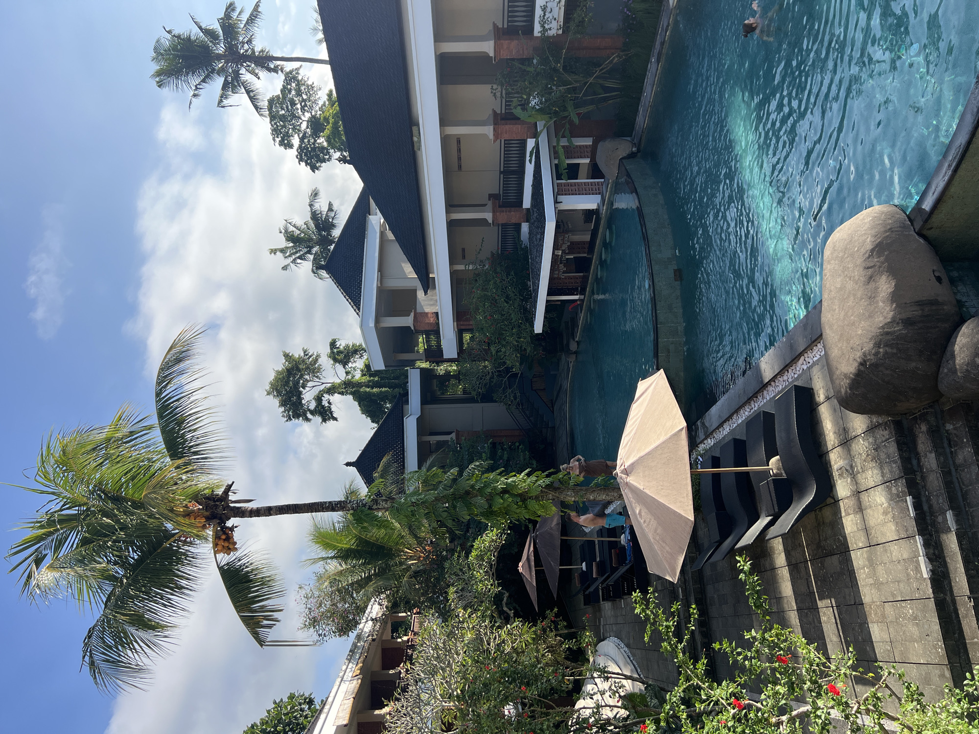 The image shows a tropical outdoor scene in Ubud District, Indonesia. The main focus is a swimming pool with clear turquoise water, bordered by stone structures and lush greenery. Adjacent to the pool, there is a shaded seating area with loungers and an umbrella. Nearby, multi-story buildings with white exteriors and dark roofs are interspersed with tropical plants, including several palm trees. The sky is blue with scattered clouds. In the pool, there is at least one person swimming.