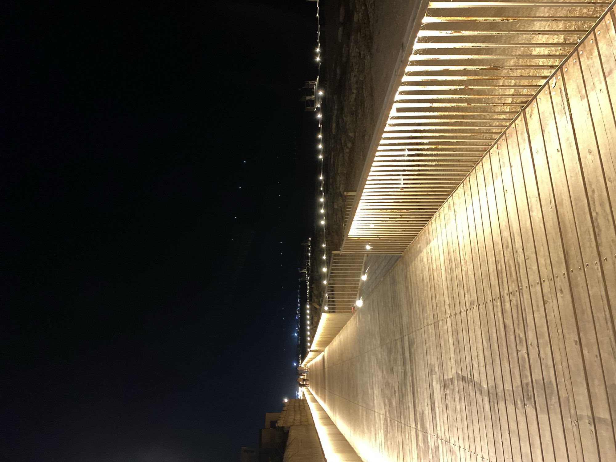 The image shows a wooden boardwalk at night, illuminated by warm lights along its length and railing. The boardwalk extends into the distance, where more lights are visible along a shoreline or landscape. A few individuals are walking on the boardwalk in the mid-ground. The sky is dark and clear. The scene depicts a well-lit promenade, likely in an outdoor public space. Given the context of Derbent, Russia, this could be a section of the Derbent embankment or a similar waterfront recreational area.