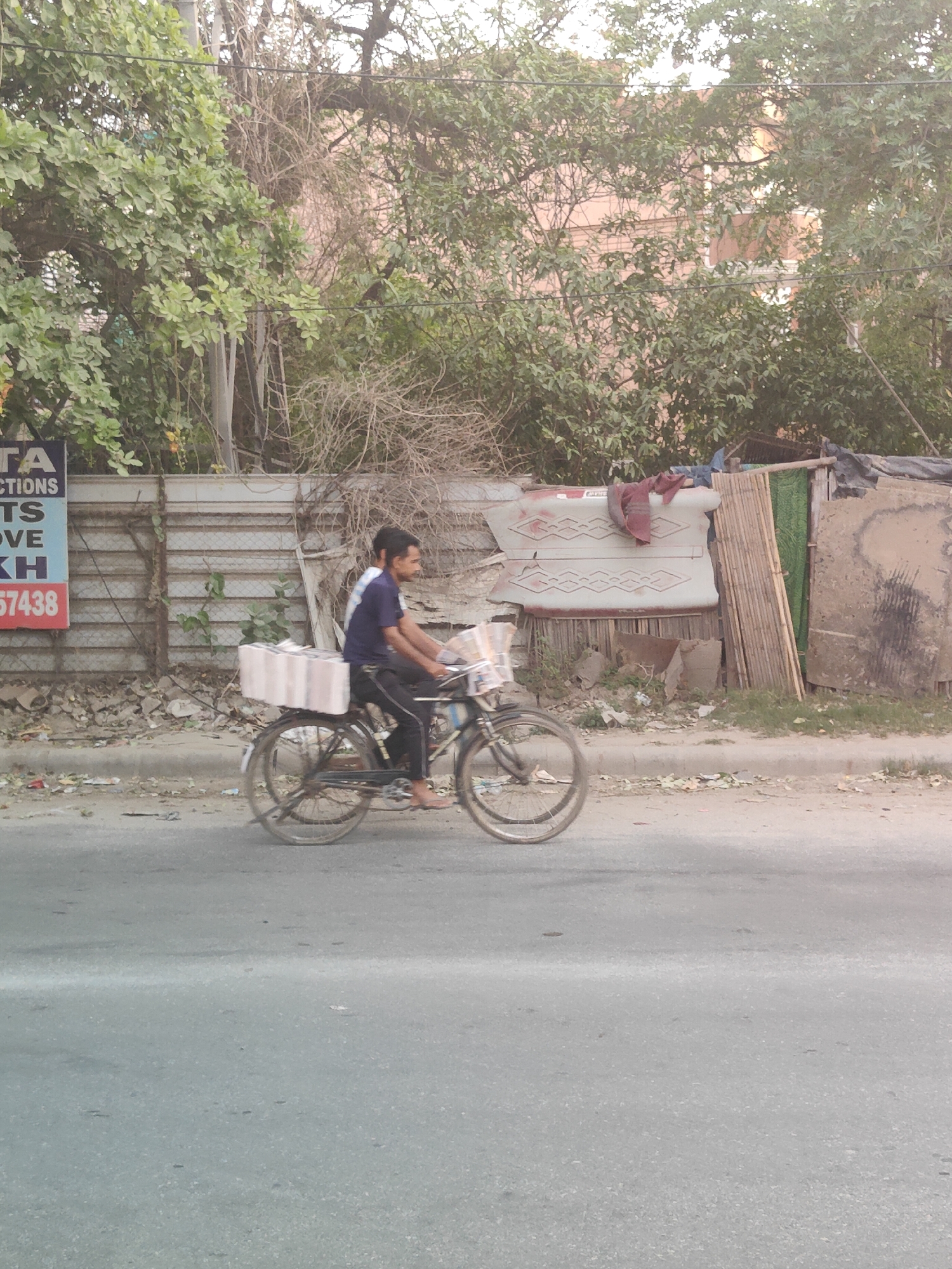 A man is riding a bicycle down a street in Gurgaon, India, with boxes loaded on the back. The man is wearing a blue t-shirt and black pants. The bicycle is a standard men's bicycle with a basket on the front and boxes on the rear rack. The boxes on the back are white with pink stripes. The street appears to be paved with asphalt and there is a sidewalk to the left of the street. Behind the sidewalk, there is a fence and some trees. The fence is made of corrugated metal and there are some posters on it. The trees are green and leafy. The sky is overcast. The overall impression is that of a busy street in a developing city.
