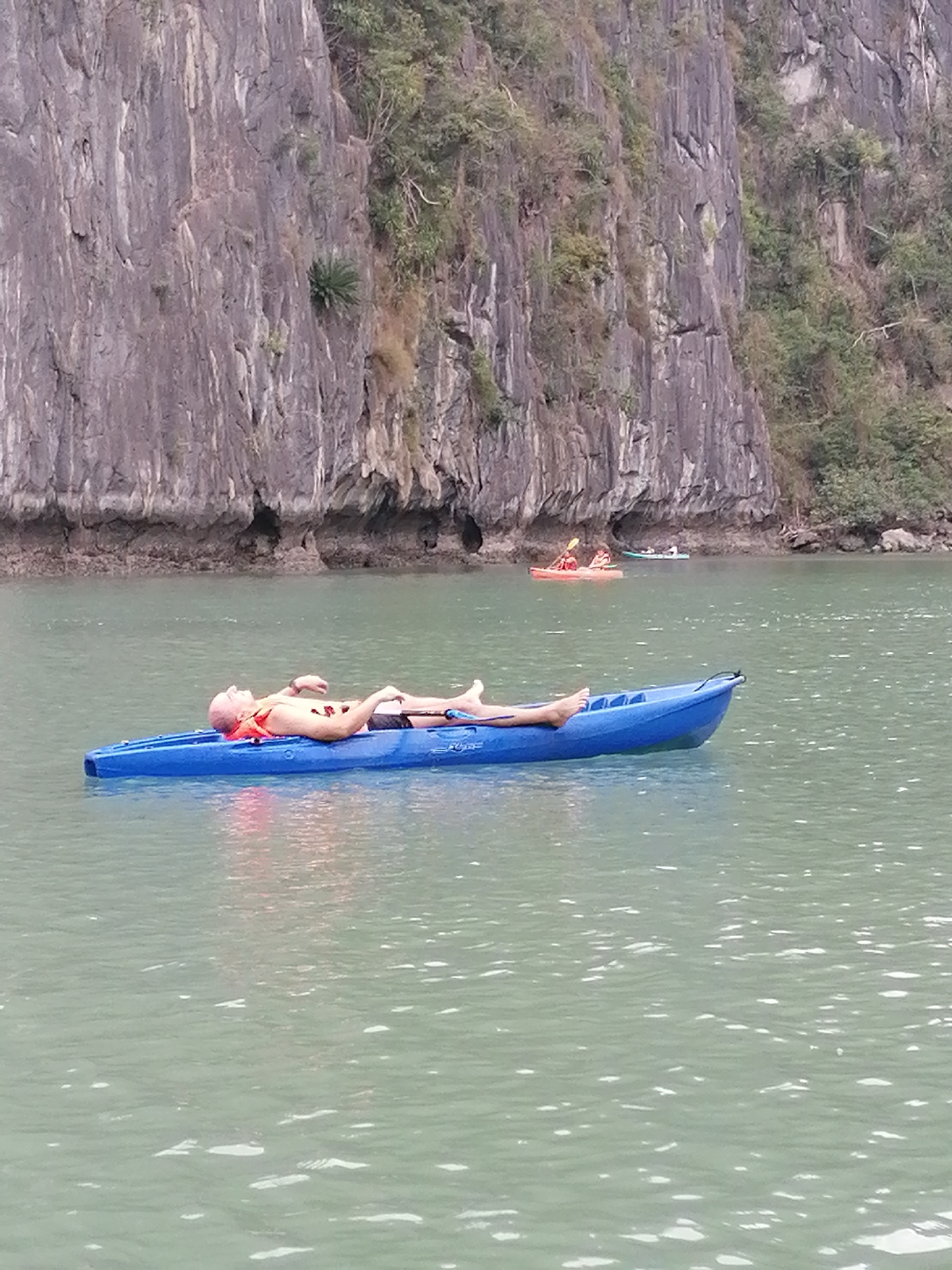 The image presents a tranquil daytime scene, characteristic of Ha Long Bay in Ha Long Ward, Vietnam, featuring towering limestone karsts emerging from calm, greenish-blue waters. The rugged cliffs in the background are primarily gray, adorned with sparse green vegetation, including small trees and shrubs.
In the foreground, a bald man with fair skin is seen relaxing in a bright blue kayak. He is lying on his back, wearing black swim shorts and a prominent orange life vest, with his arms gently bent and legs extended. The kayak has the brand name "Perception" visible on its side. His relaxed posture suggests he is either sunbathing or simply enjoying the peaceful environment.
Further back on the water, other kayakers are visible. To the right of the man, a red kayak with one or two individuals actively paddling can be seen. Beyond them, a lighter-colored kayak with another person is present, and a very distant boat or kayak is faintly discernible near the base of the cliffs.
The weather appears clear and calm, contributing to the smooth surface of the water. The diffused lighting suggests it is daytime, possibly late morning or early afternoon, creating a serene and picturesque setting for recreational activity on the water.
