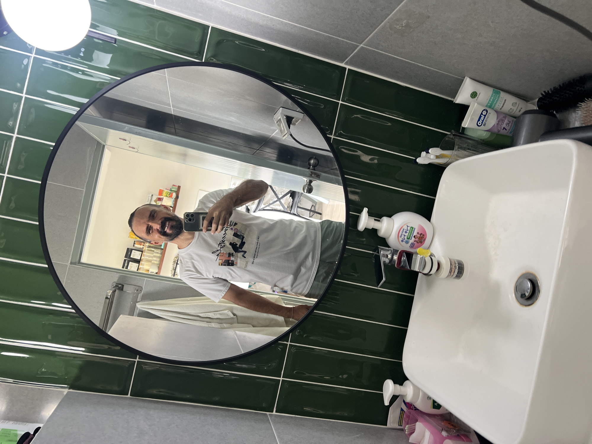 Here is a forensic description of the image:
**Scene:** Interior of a bathroom, seemingly in a private residence.
**Location:** Likely in the vicinity of บ้านบางรักษ์, Thailand, based on the presence of Thai language packaging on some of the toiletries.
**People:** One person is visible as a reflection in a round mirror. He is a bearded male, holding a mobile phone to take the picture. He is wearing a white t-shirt with a graphic design.
**Objects:**
* A round mirror mounted on a green tiled wall.
* A white rectangular washbasin.
* Various toiletries including toothpaste (Oral-B), soap bottles (one with Thai script).
* A grey-tiled counter.
* A towel rack and a towel are visible in the background reflection.
**Actions/Interactions:**
* The man is taking a selfie in the bathroom mirror.
**Environment:**
* The walls are partially covered in green, rectangular tiles, and partially in gray tiles.
* The room appears to be well-lit, with a light fixture visible above the mirror.
* The overall environment suggests a modern, clean bathroom.