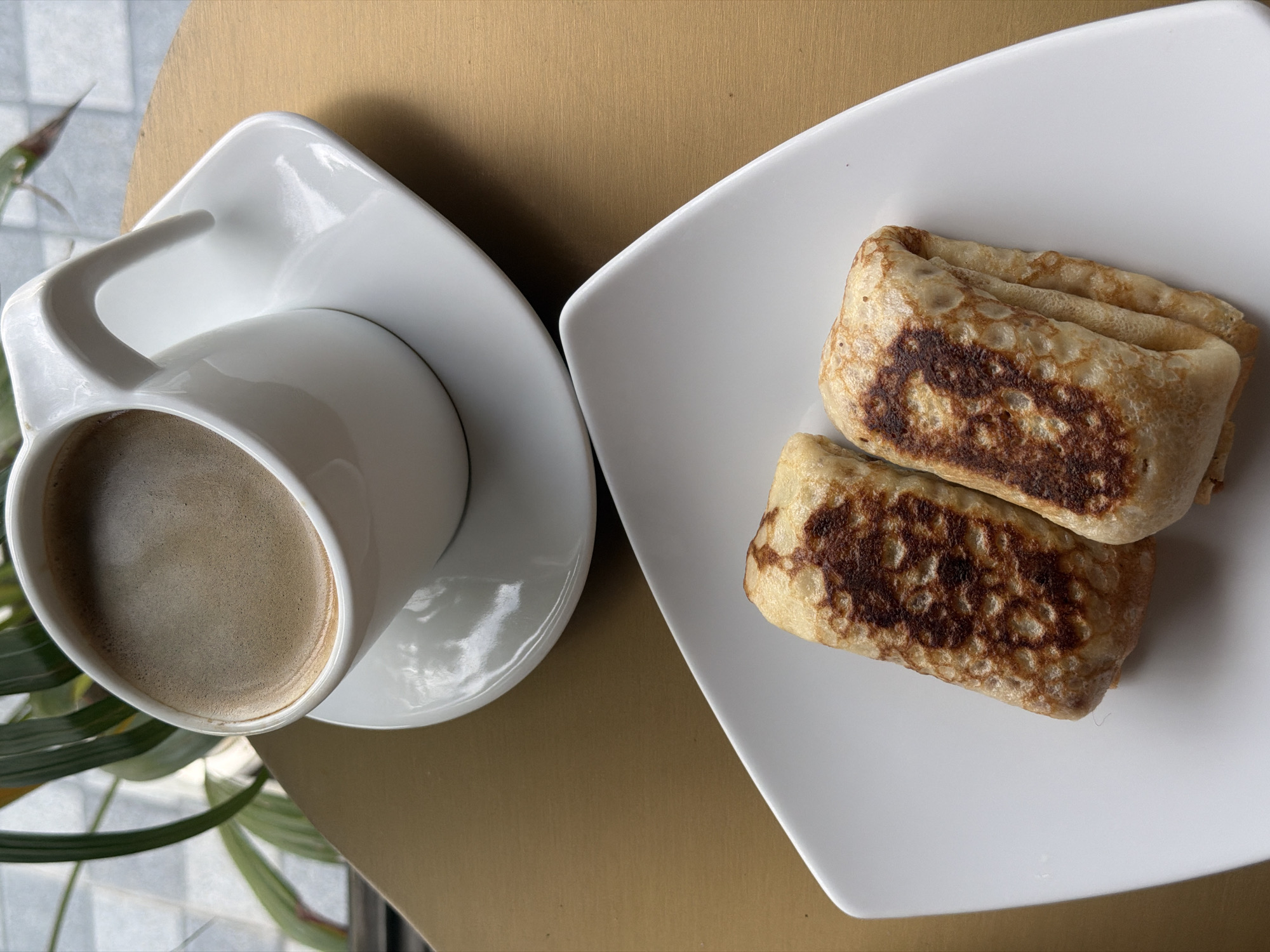 A white ceramic cup filled with coffee sits on a white saucer, which is placed on a golden-brown surface. To the left of the cup, green leaves of a plant are visible, potentially indicating an indoor or semi-outdoor setting. In the foreground, on a white plate, are two folded crepes or thin pancakes. The crepes have a golden-brown color with darker browned spots, suggesting they have been cooked. The image is a still life, showcasing a breakfast or snack. The location in Ko Samui District, Thailand, is not discernible from the visual elements of the image itself.