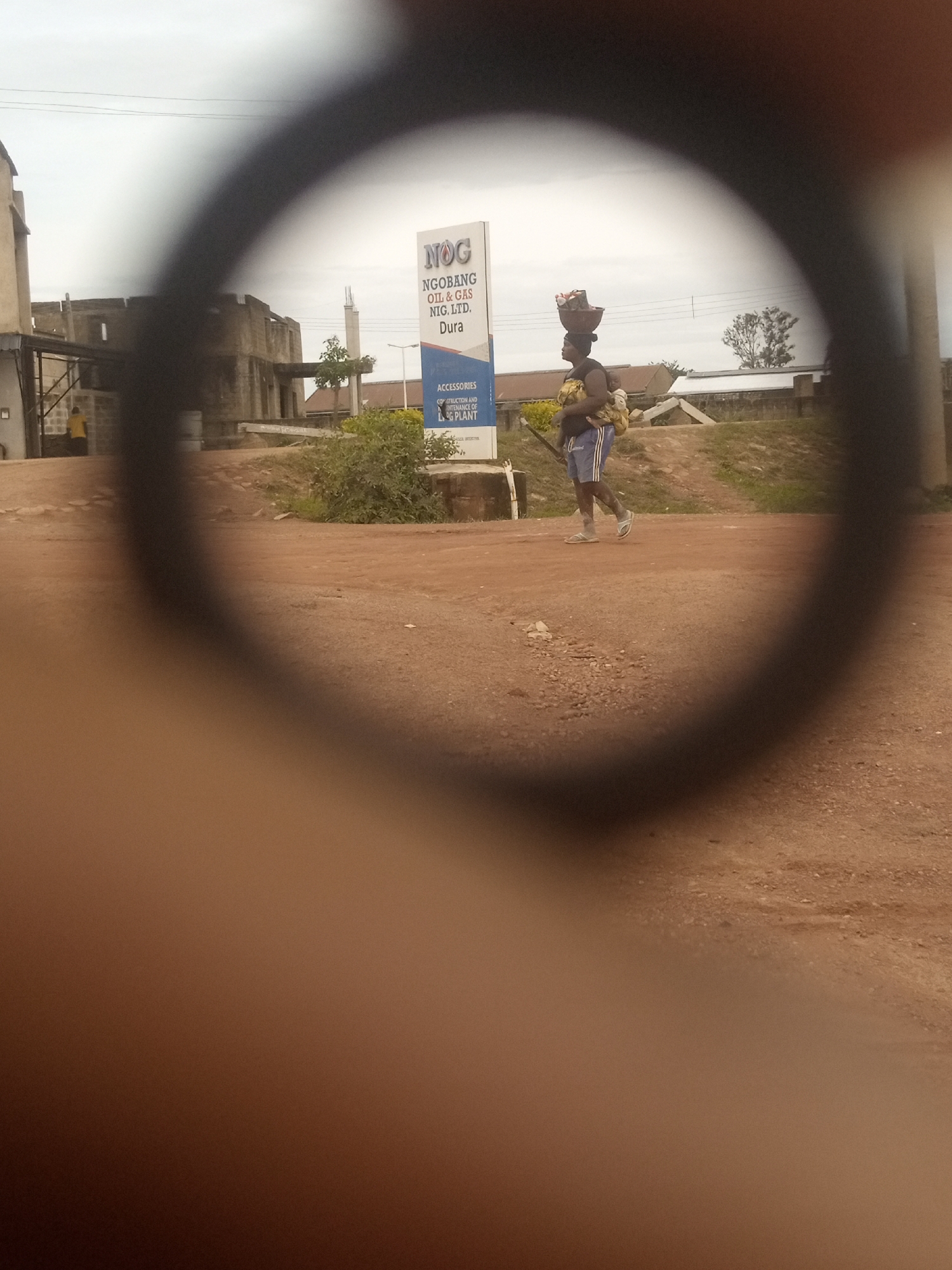 The image captures an outdoor scene in Kwang, Nigeria, viewed through a circular, blurred lens, likely from a camera or optical device. In the foreground, obscuring a large portion of the frame, is a dark, blurred ring. The background reveals a street scene.
The focal point of the image is a person walking, carrying items on their head and holding a child or another item against their body. The person is wearing blue shorts and a dark top. They are walking on a dirt road, which appears to be uneven with small stones and patches of dry earth.
To the left of the person is a tall, white sign with text that is partially visible and difficult to read clearly due to the overall blur. It appears to be an advertisement or informational sign, possibly for a company or service. Behind the sign and the person, there are low-rise buildings and vegetation. The sky is overcast, suggesting a cloudy day. The environment appears to be a rural or semi-urban setting within Kwang, Nigeria.