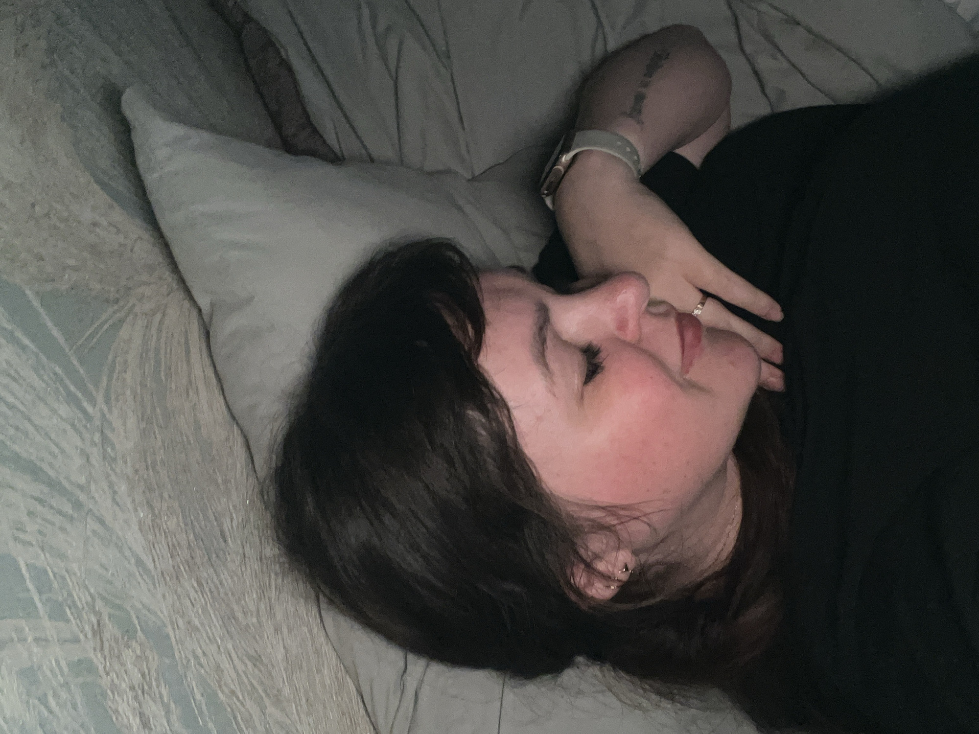 A close-up, dimly lit image shows a person, likely female, lying on their left side on a bed. Their eyes are closed, suggesting they are asleep or resting peacefully. The person has dark hair, which is styled with bangs and frames their face, and fair skin with a reddish flush on the visible cheek.
Their head rests on a light-colored pillow, and they are covered by a light blue and gray patterned blanket or duvet. The person is wearing a dark top or shirt. Their left arm is bent, with the hand resting near their face. A light-colored smartwatch is visible on their left wrist, and a ring with a stone adorns their left ring finger. A small stud or hoop earring is in their left ear. A tattoo with what appears to be cursive script is visible on their upper left arm, but the text is illegible. The subdued lighting indicates that it may be nighttime or a very dimly lit room. The scene conveys a sense of tranquility and rest. The location context for this media file is Noviy, Russia.