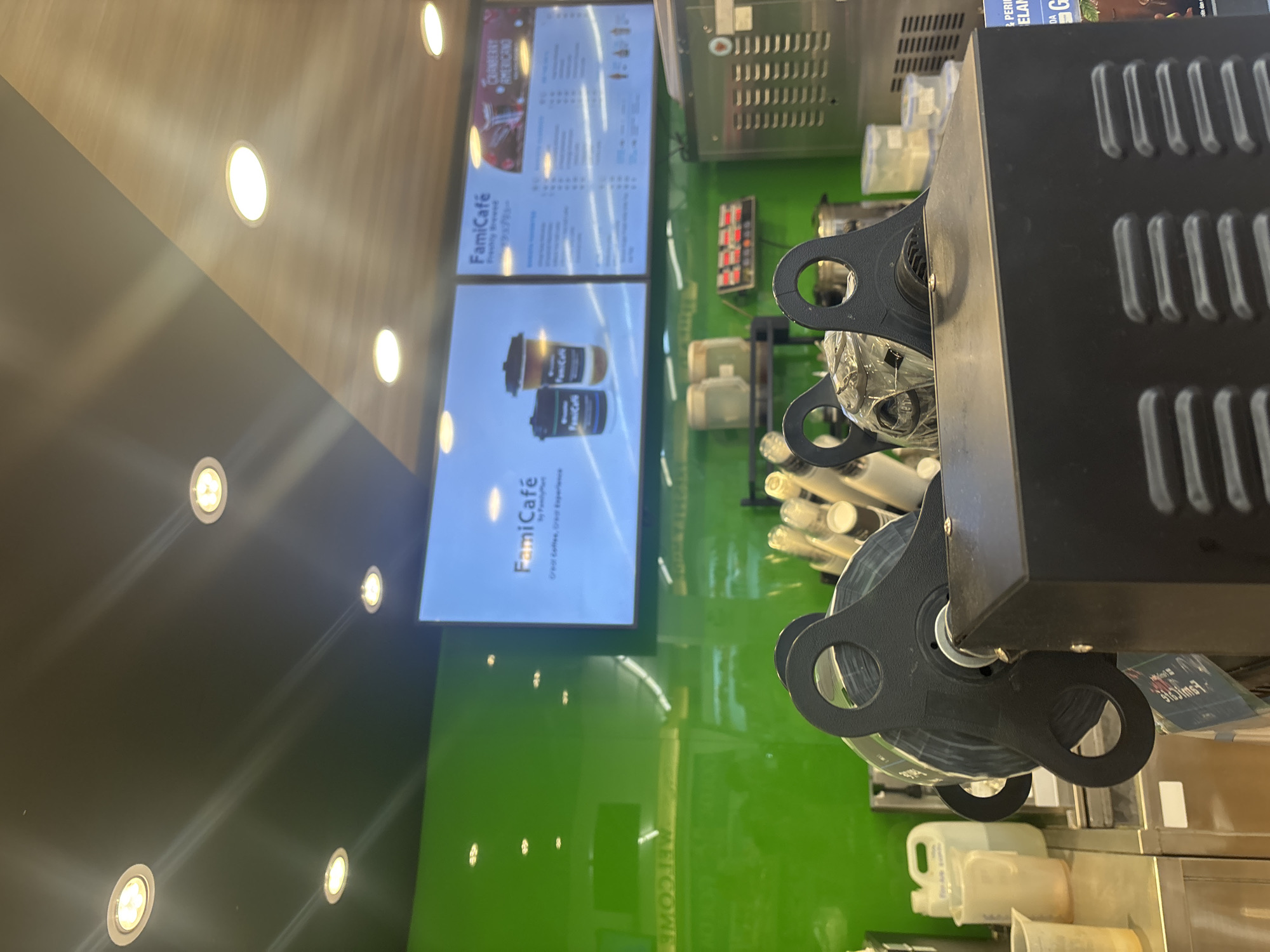 The image shows an interior view of a FamiCafe counter area, likely located in East Jakarta, Indonesia. In the foreground, a black machine with large, circular handles is prominently displayed. Behind it, a bright green wall serves as a backdrop. Above the green wall, a large digital display shows promotional content for FamiCafe, featuring images of coffee cups and text such as "FamiCafe" and "Great Coffee, Great Experience." Another section of the display lists menu items, including "CRANBERRY AMERICANO." Various cafe equipment, including what appear to be coffee bean dispensers and beverage containers, are visible on the counter and shelves. The lighting suggests an indoor environment with overhead spotlights.