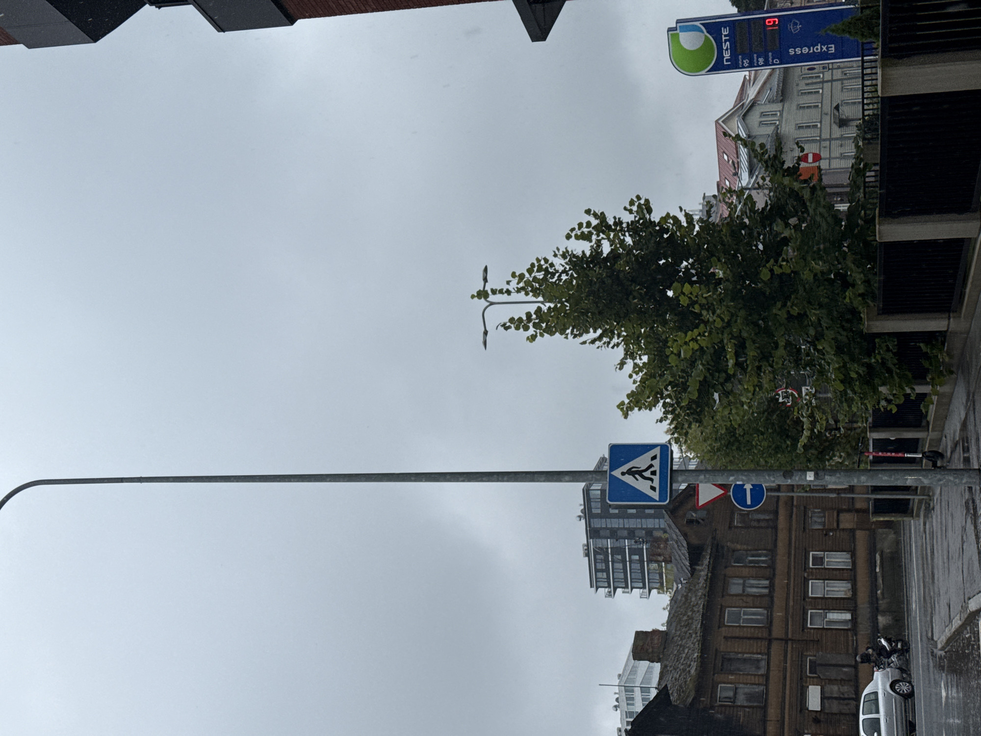The image captures a street scene in Tallinn, Estonia, on an overcast day, likely with light rain. A prominent tall, thin lamppost stands to the left of the frame. Below it, a pedestrian crossing sign with a blue square and a white silhouette of a person walking is visible, accompanied by a red and white triangular yield sign and a blue circular sign with a white arrow pointing right.
To the left, a two-story wooden building with a weathered facade and multiple windows lines the street. A silver car and a person on a motorbike are parked on the wet asphalt road in front of it. Further back, modern apartment buildings rise above the older structures.
A lush green tree with dense foliage is positioned in the center-right of the image, partially obscuring the view of other buildings. To the far right, a Neste gas station sign with the company's logo and pricing information for different fuel types is displayed. The price for "Express" fuel is shown as 1.9. A black metal fence runs along the bottom of the image, separating the street from what appears to be a walkway or private area. The sky is uniformly grey, suggesting cloudy weather.